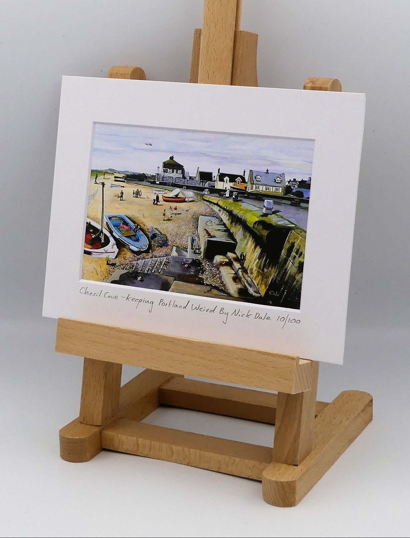 Image of a Small Art Print featuring a copy of the painting 'Chesil Cove - Keeping Portland Weird' by Nick Dale. The print depicts the end of Chesil Beach where it meets Portland at Chesil Cove. In the scene the size of the people is not affected by their distance from the foreground, hence making their appearance slightly surreal. The print is situated on a small easel.