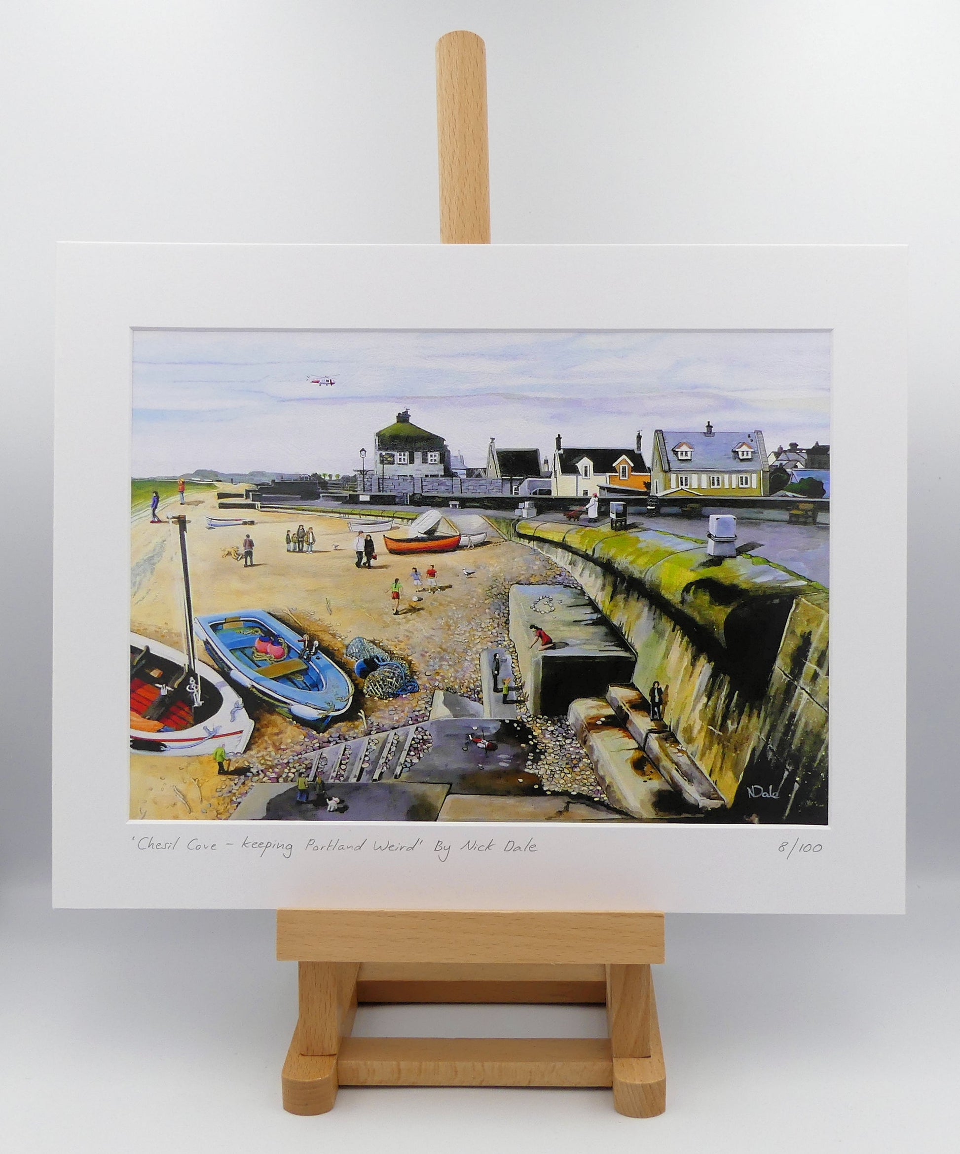 Image of a Medium Art Print featuring a copy of the painting 'Chesil Cove - Keeping Portland Weird' by Nick Dale. The print depicts the end of Chesil Beach where it meets Portland at Chesil Cove. In the scene the size of the people is not affected by their distance from the foreground, hence making their appearance slightly surreal. The print is situated on a small easel.