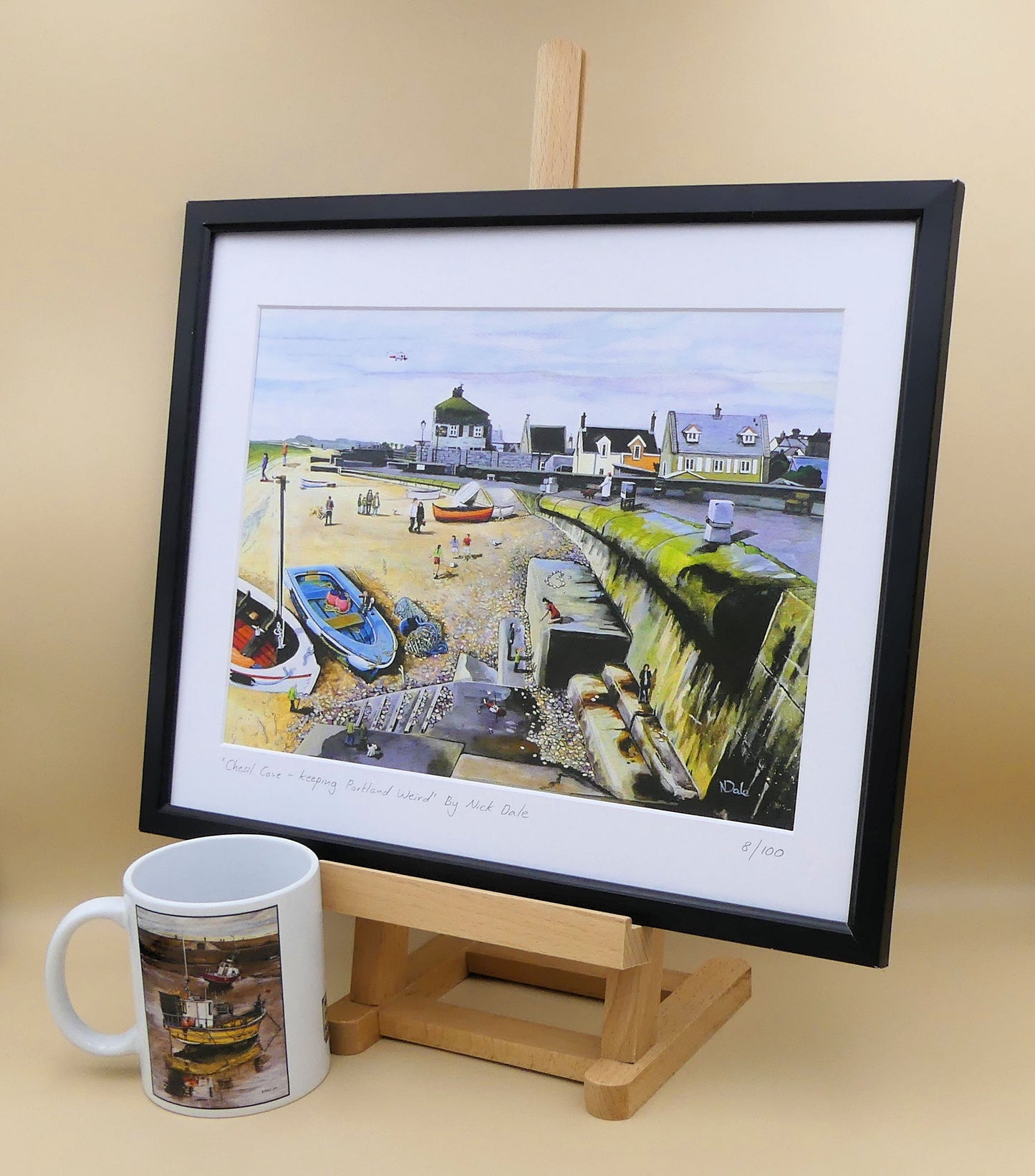 Image of a Medium Art Print featuring a copy of the painting 'Chesil Cove - Keeping Portland Weird' by Nick Dale. The print depicts the end of Chesil Beach where it meets Portland at Chesil Cove. In the scene the size of the people is not affected by their distance from the foreground, hence making their appearance slightly surreal. The print is situated on a small easel in a black frame next to a mug.