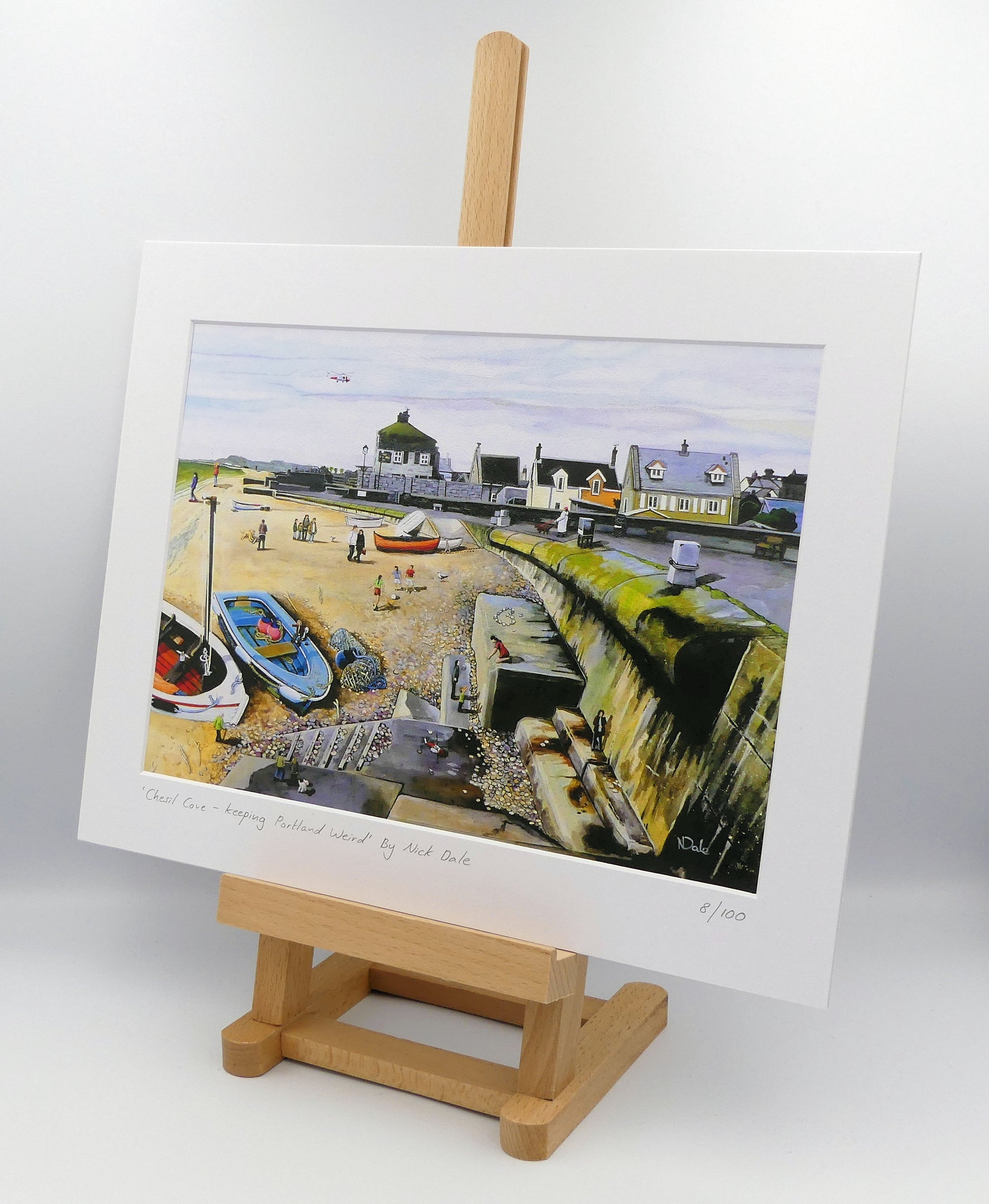Image of a Medium Art Print featuring a copy of the painting 'Chesil Cove - Keeping Portland Weird' by Nick Dale. The print depicts the end of Chesil Beach where it meets Portland at Chesil Cove. In the scene the size of the people is not affected by their distance from the foreground, hence making their appearance slightly surreal. The print is situated on a small easel.