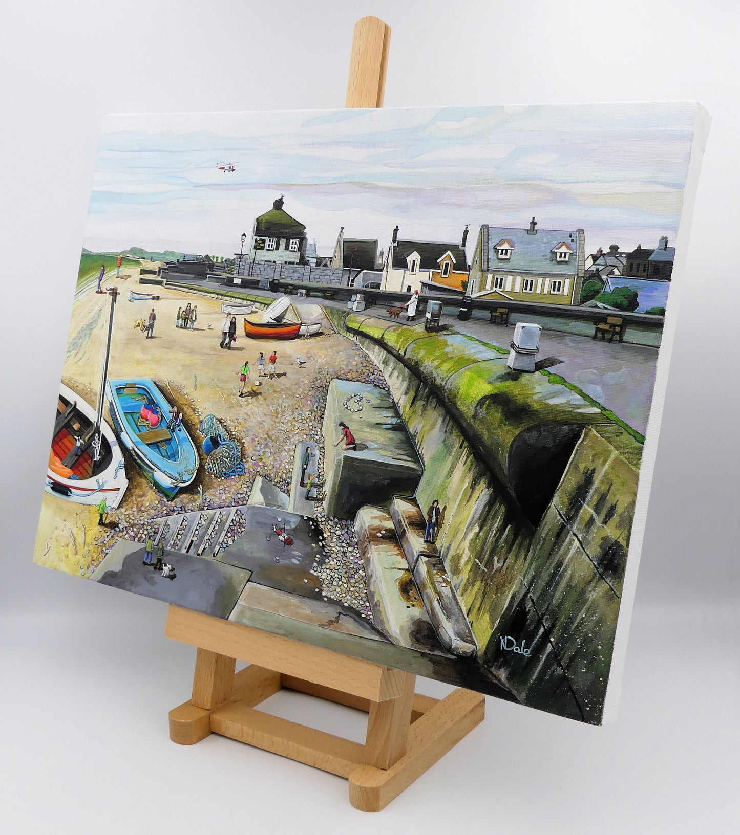 Image of the original painting 'Chesil Cove - Keeping Portland Weird' by Nick Dale on a small easel.  The scene depicts the end of Chesil Beach where it meets Portland at Chesil Cove. In the painting the size of the people is not affected by their distance from the foreground, hence making their appearance slightly surreal. 