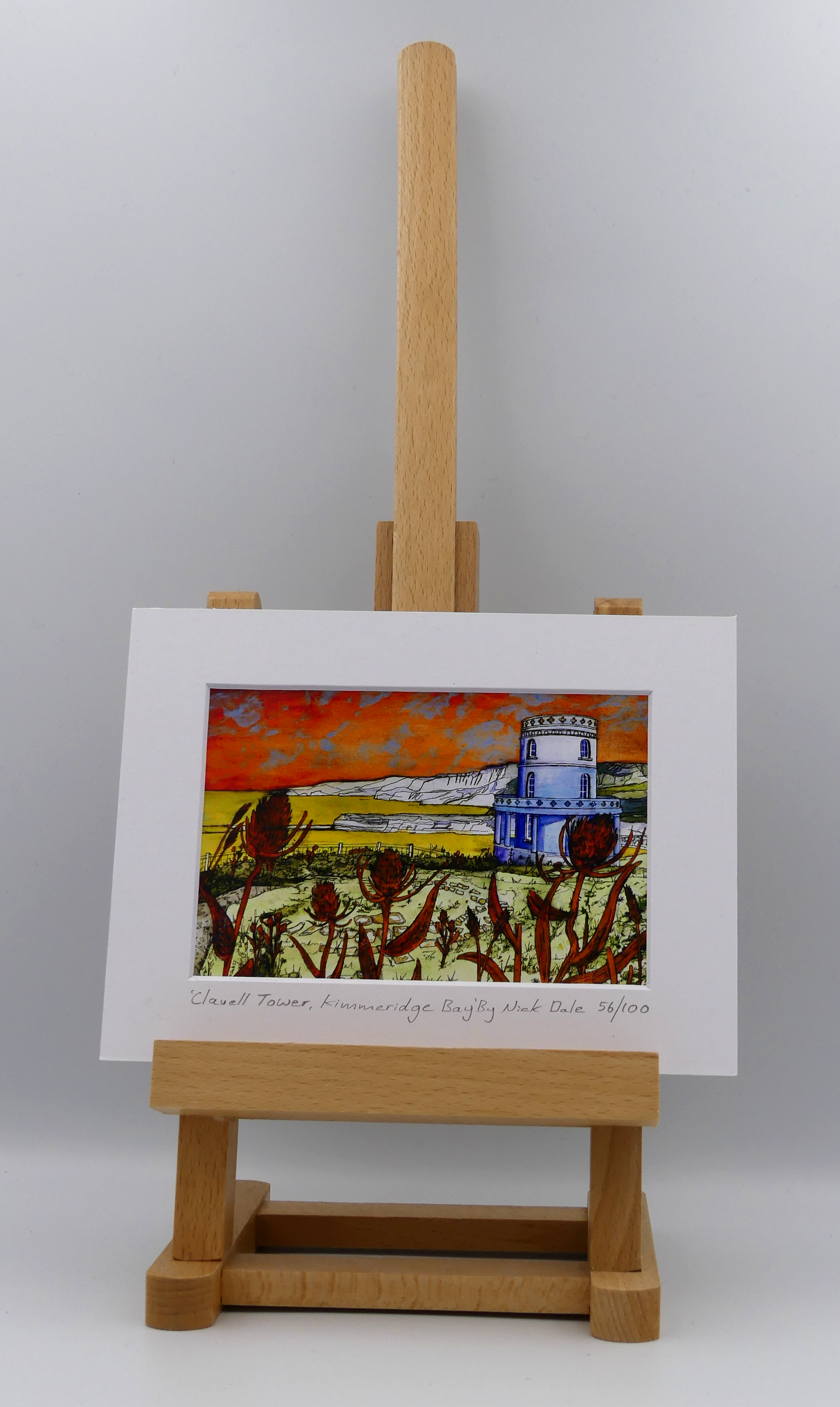 "Image of a Small Art Print featuring a copy of the painting 'Clavell Tower, Kimmeridge Bay' by Nick Dale. The scene depicts Clavell Tower (which is also known as the Clavell Folly or the Kimmeridge Tower) situated on the cliffs overlooking Kimmeridge Bay on the Isle of Purbeck, Dorset. 
The tower; which was built in 1830, is seen in its current position with the original footings visible in the foreground after the tower was moved further back from the cliff in 2006. The print is situated on a small easel.