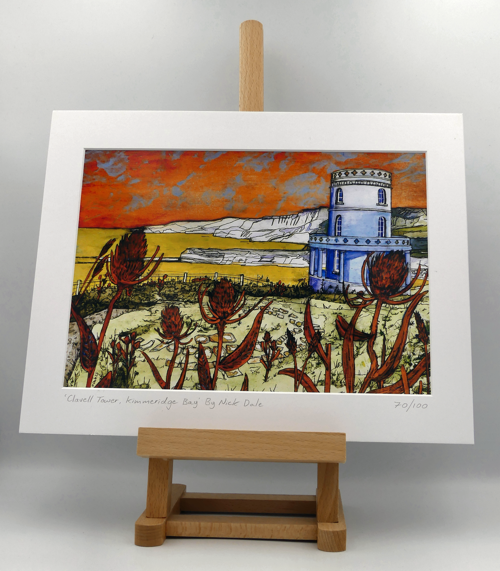 Image of a Medium Art Print featuring a copy of the painting 'Clavell Tower, Kimmeridge Bay' by Nick Dale. The scene depicts Clavell Tower (which is also known as the Clavell Folly or the Kimmeridge Tower) situated on the cliffs overlooking Kimmeridge Bay on the Isle of Purbeck, Dorset. 
The tower; which was built in 1830, is seen in its current position with the original footings visible in the foreground after the tower was moved further back from the cliff in 2006. The print is situated on a small easel.