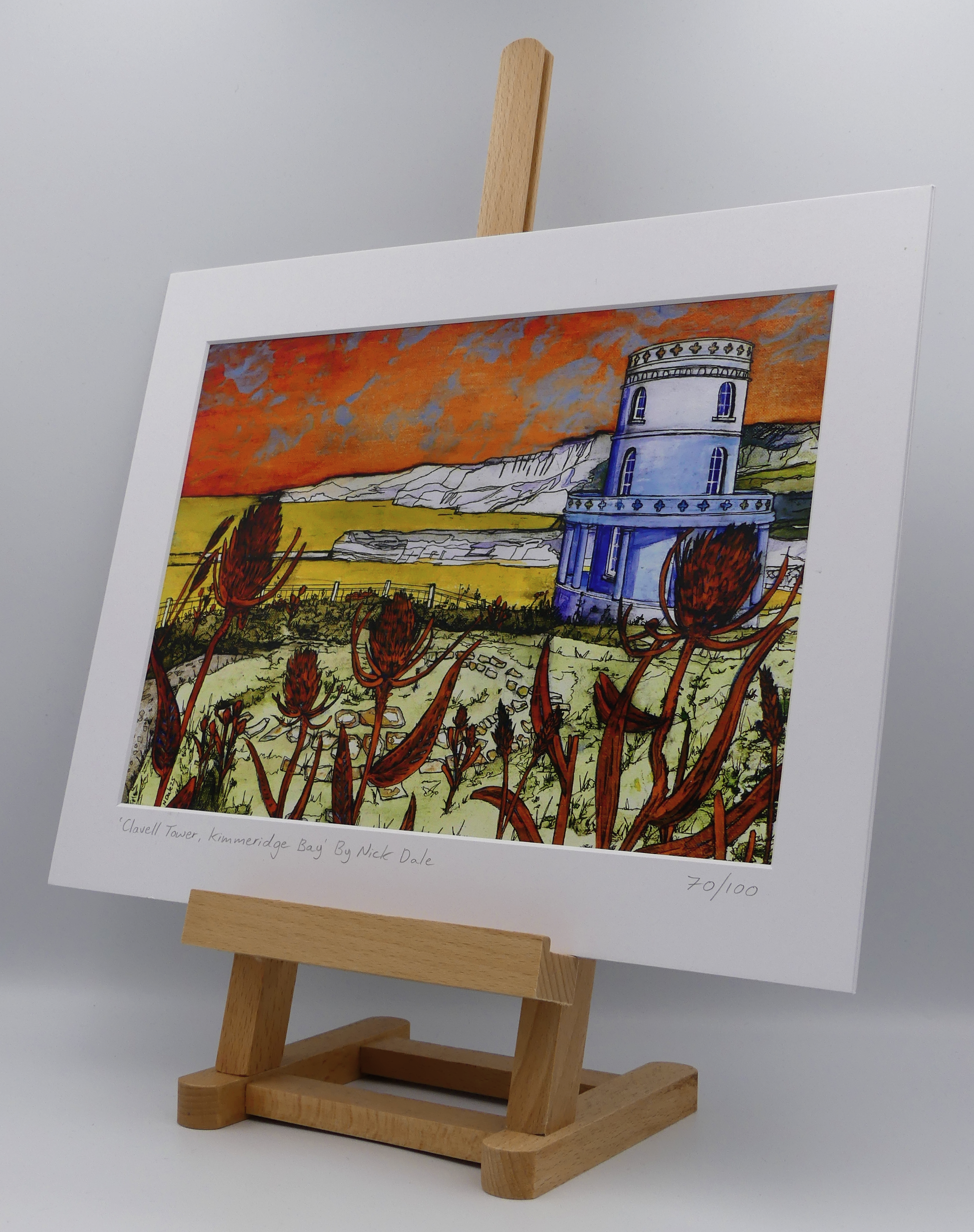 Image of a Medium Art Print featuring a copy of the painting 'Clavell Tower, Kimmeridge Bay' by Nick Dale. The scene depicts Clavell Tower (which is also known as the Clavell Folly or the Kimmeridge Tower) situated on the cliffs overlooking Kimmeridge Bay on the Isle of Purbeck, Dorset. 
The tower; which was built in 1830, is seen in its current position with the original footings visible in the foreground after the tower was moved further back from the cliff in 2006. The print is situated on a small easel.
