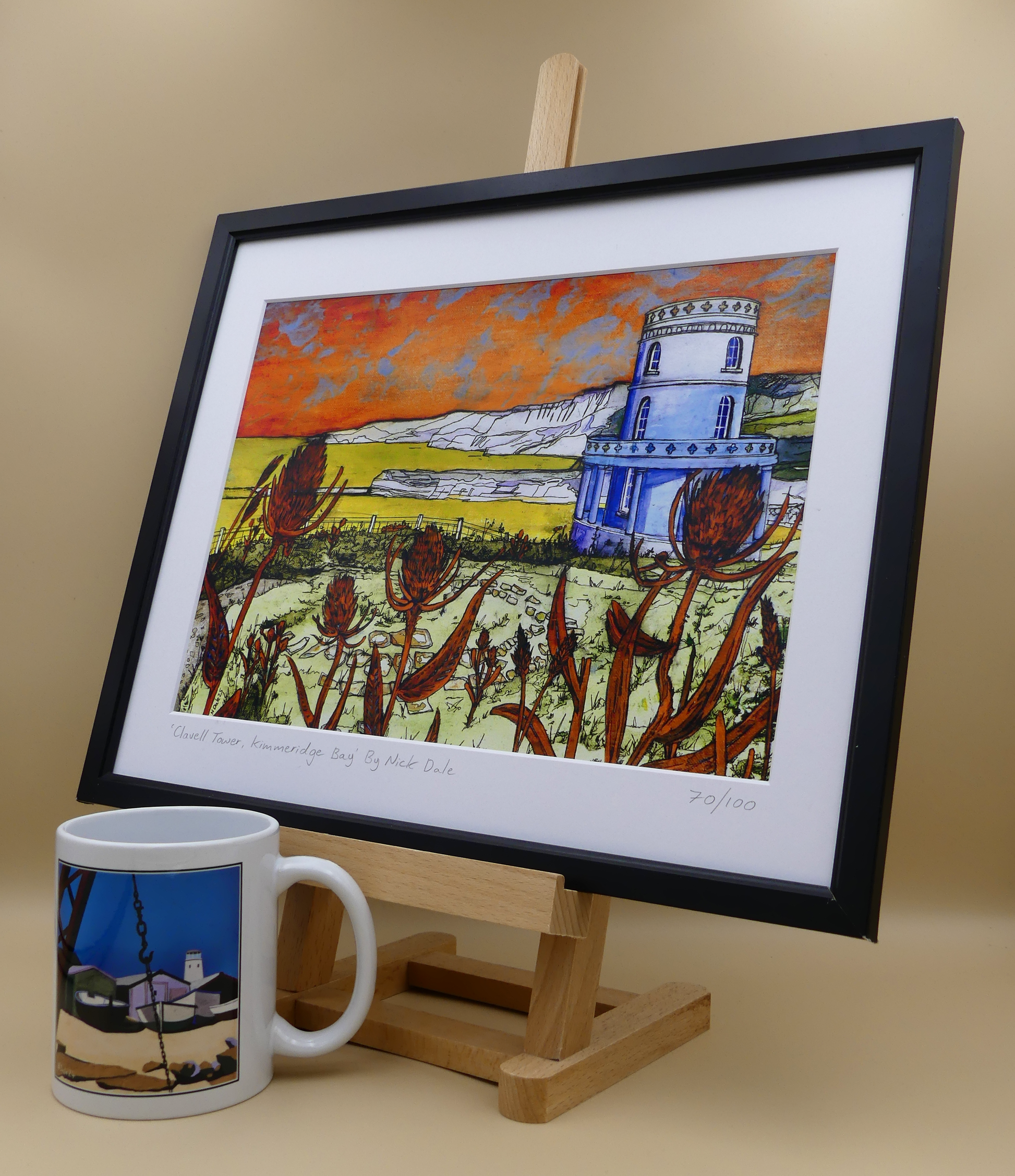 Image of a Medium Art Print featuring a copy of the painting 'Clavell Tower, Kimmeridge Bay' by Nick Dale. The scene depicts Clavell Tower situated on the cliffs overlooking Kimmeridge Bay on the Isle of Purbeck, Dorset. 
The tower; which was built in 1830, is seen in its current position with the original footings visible in the foreground after the tower was moved further back from the cliff in 2006. The print is situated on a small easel in a black frame next to a mug.