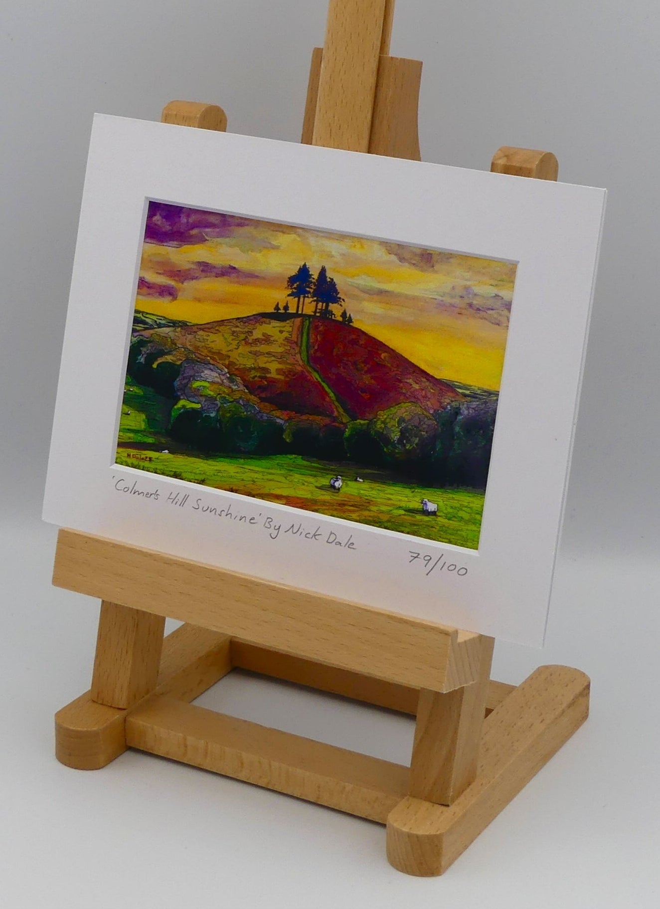 Image of a Small Art Print featuring a copy of the painting 'Colmer's Hill Sunshine' by Nick Dale. The print depicts the prominent sandstone conical hill near the village of Symondsbury in West Dorset. The hill is topped with pine trees to commemorate those who died in World War I. The print is situated on a small easel.