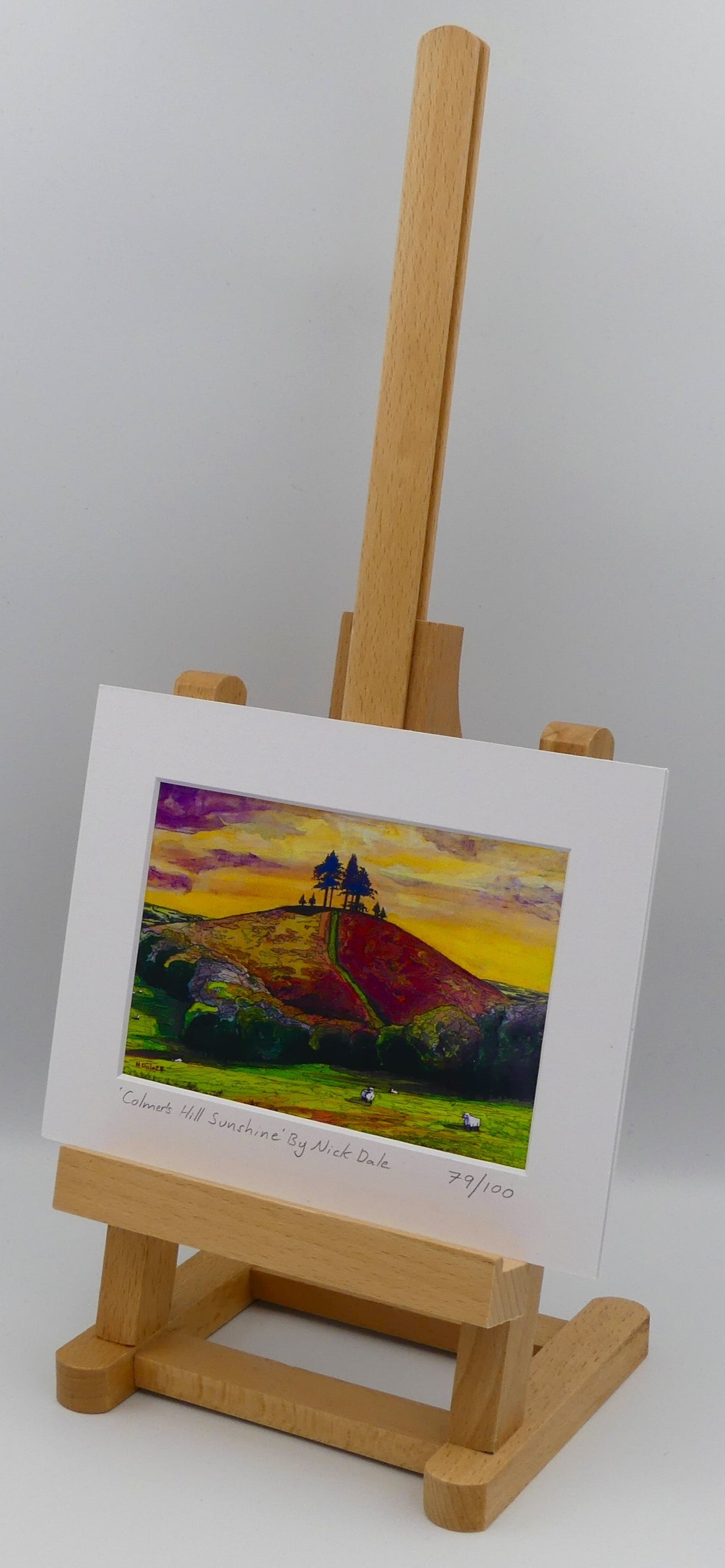 Image of a Small Art Print featuring a copy of the painting 'Colmer's Hill Sunshine' by Nick Dale. The print depicts the prominent sandstone conical hill near the village of Symondsbury in West Dorset. The hill is topped with pine trees to commemorate those who died in World War I. The print is situated on a small easel.