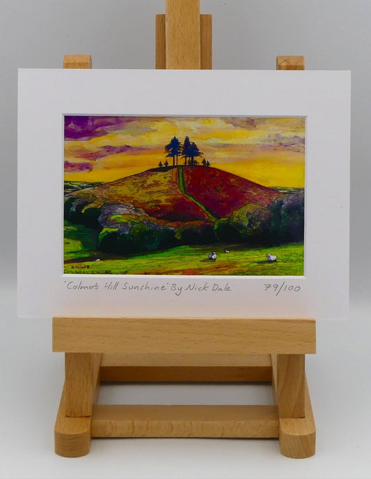Image of a Small Art Print featuring a copy of the painting 'Colmer's Hill Sunshine' by Nick Dale. The print depicts the prominent sandstone conical hill near the village of Symondsbury in West Dorset. The hill is topped with pine trees to commemorate those who died in World War I.  The print is situated on a small easel.