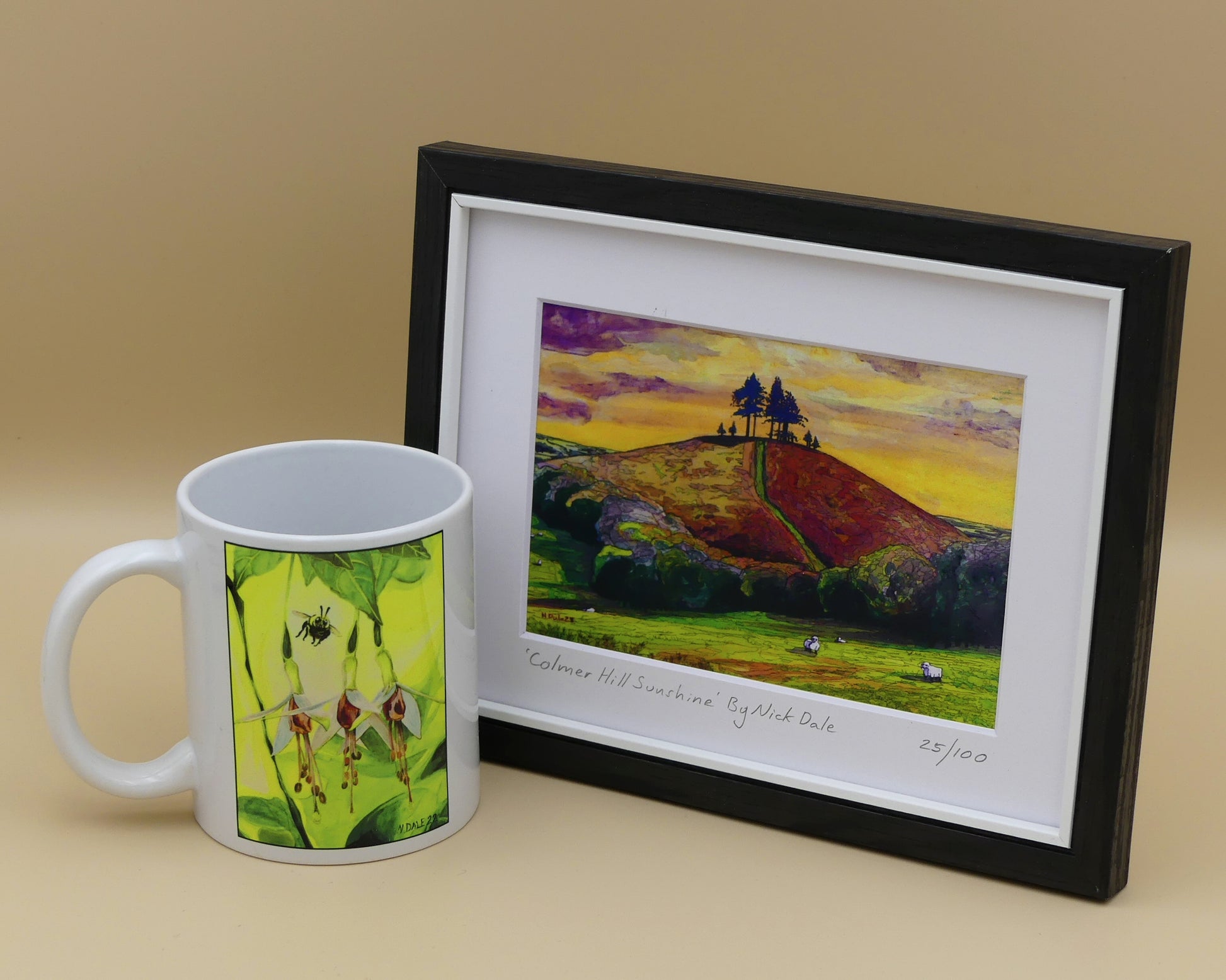 Image of a Small Art Print featuring a copy of the painting 'Colmer's Hill Sunshine' by Nick Dale. The print depicts the prominent sandstone conical hill near the village of Symondsbury in West Dorset. The hill is topped with pine trees to commemorate those who died in World War I. The print is situated in a black frame next to a mug.