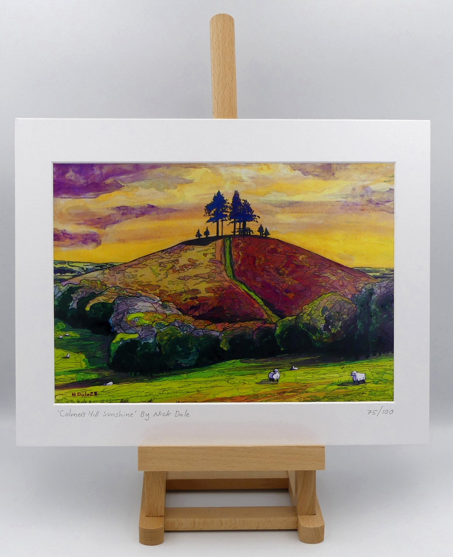 Image of a Medium Art Print featuring a copy of the painting 'Colmer's Hill Sunshine' by Nick Dale. The print depicts the prominent sandstone conical hill near the village of Symondsbury in West Dorset. The hill is topped with pine trees to commemorate those who died in World War I. The print is situated on a small easel.