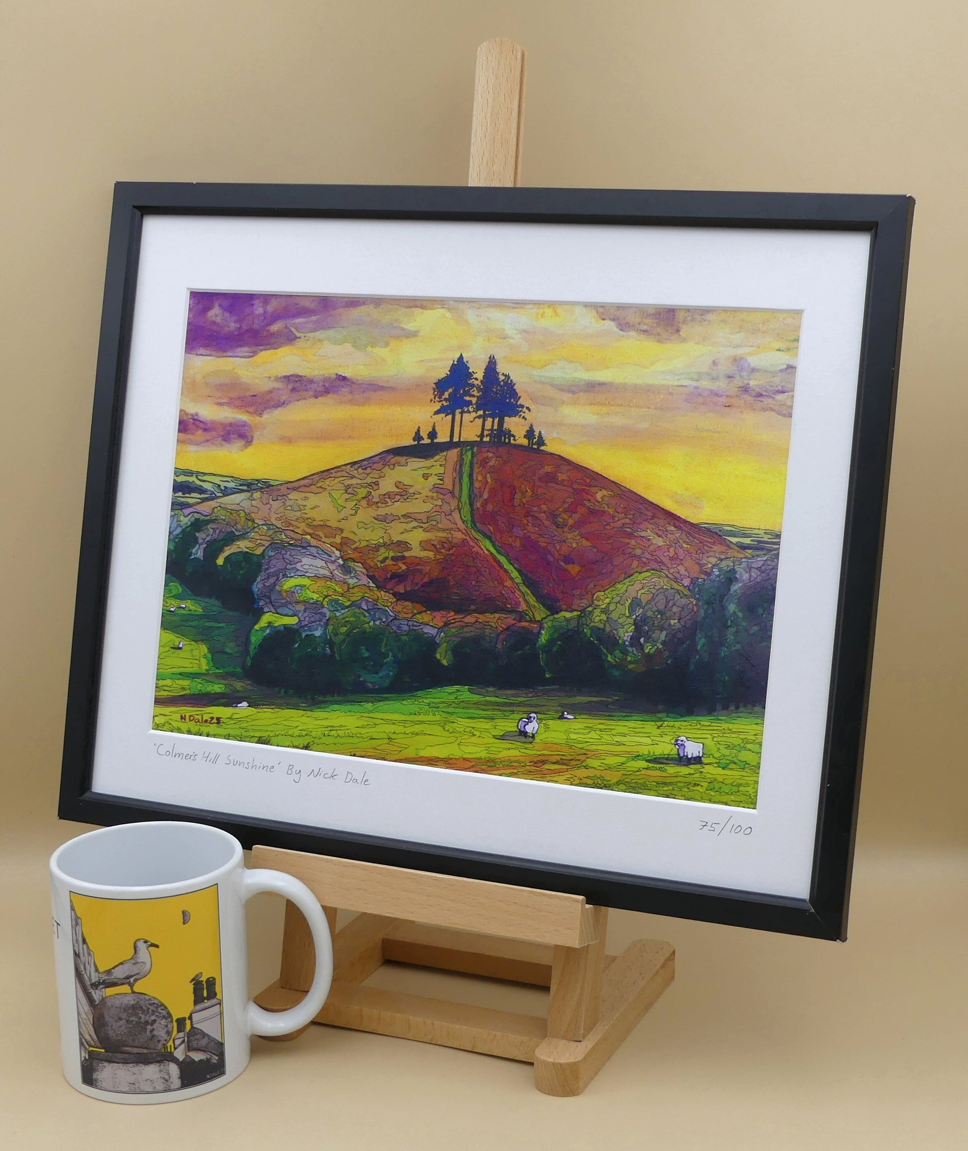 Image of a Medium Art Print featuring a copy of the painting 'Colmer's Hill Sunshine' by Nick Dale. The print depicts the prominent sandstone conical hill near the village of Symondsbury in West Dorset. The hill is topped with pine trees to commemorate those who died in World War I. The print is situated on a small easel in a black frame next to a mug.