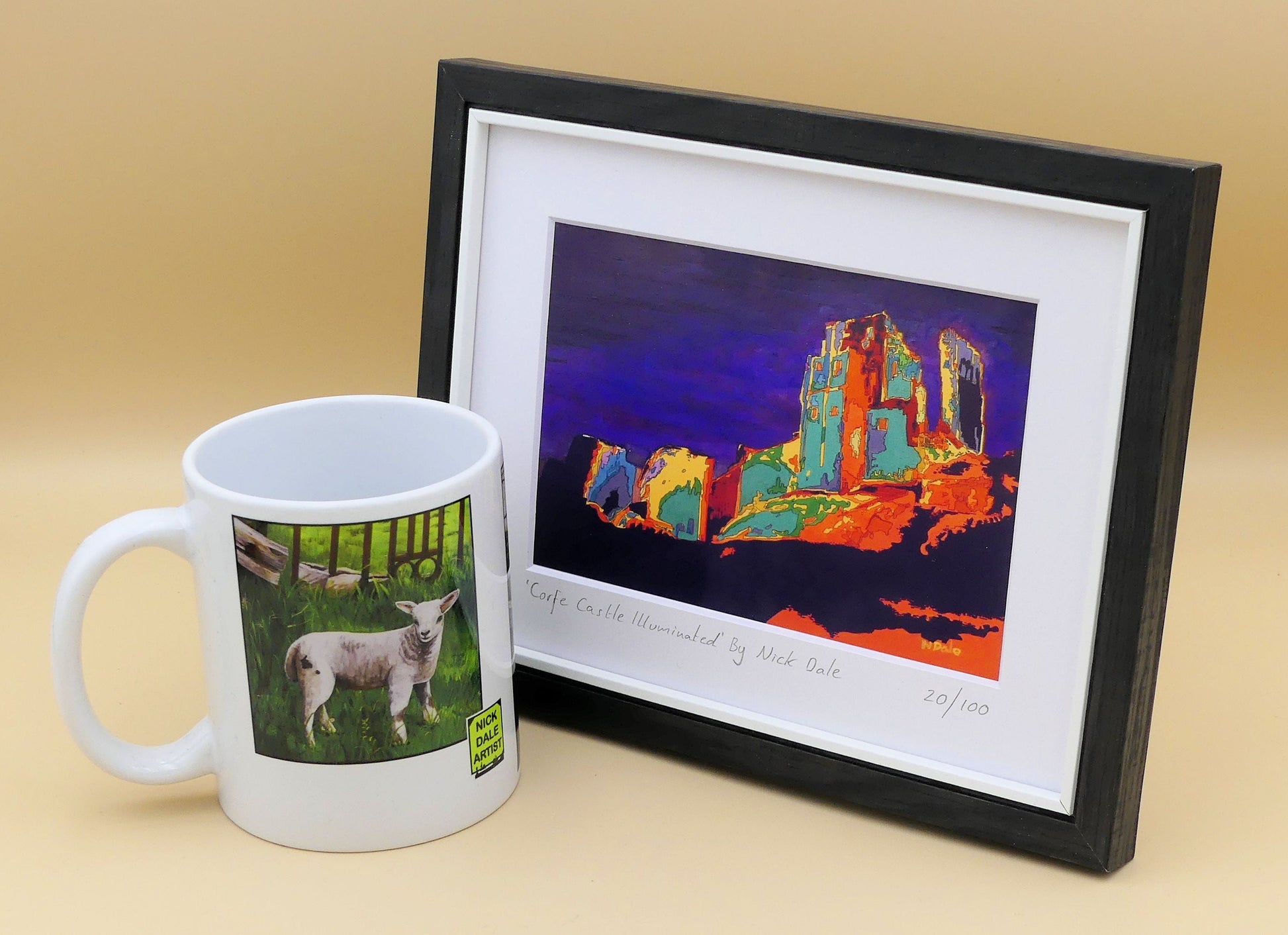 Image of a Small Art Print featuring a copy of the painting 'Corfe Castle Illuminated' by Nick Dale. The print depicts a view of the ruins of Corfe Castle at night from within the outer bailey; with the south-west gatehouse on the left and the keep on top of the central hilltop on the right. The print is situated in a black frame next to a mug.