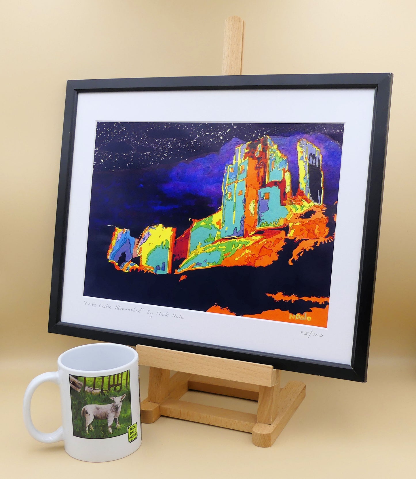 Image of a Medium Art Print featuring a copy of the painting 'Corfe Castle Illuminated' by Nick Dale. The print depicts a view of the ruins of Corfe Castle at night from within the outer bailey; with the south-west gatehouse on the left and the keep on top of the central hilltop on the right. The print is situated on a small easel in a black frame next to a mug.