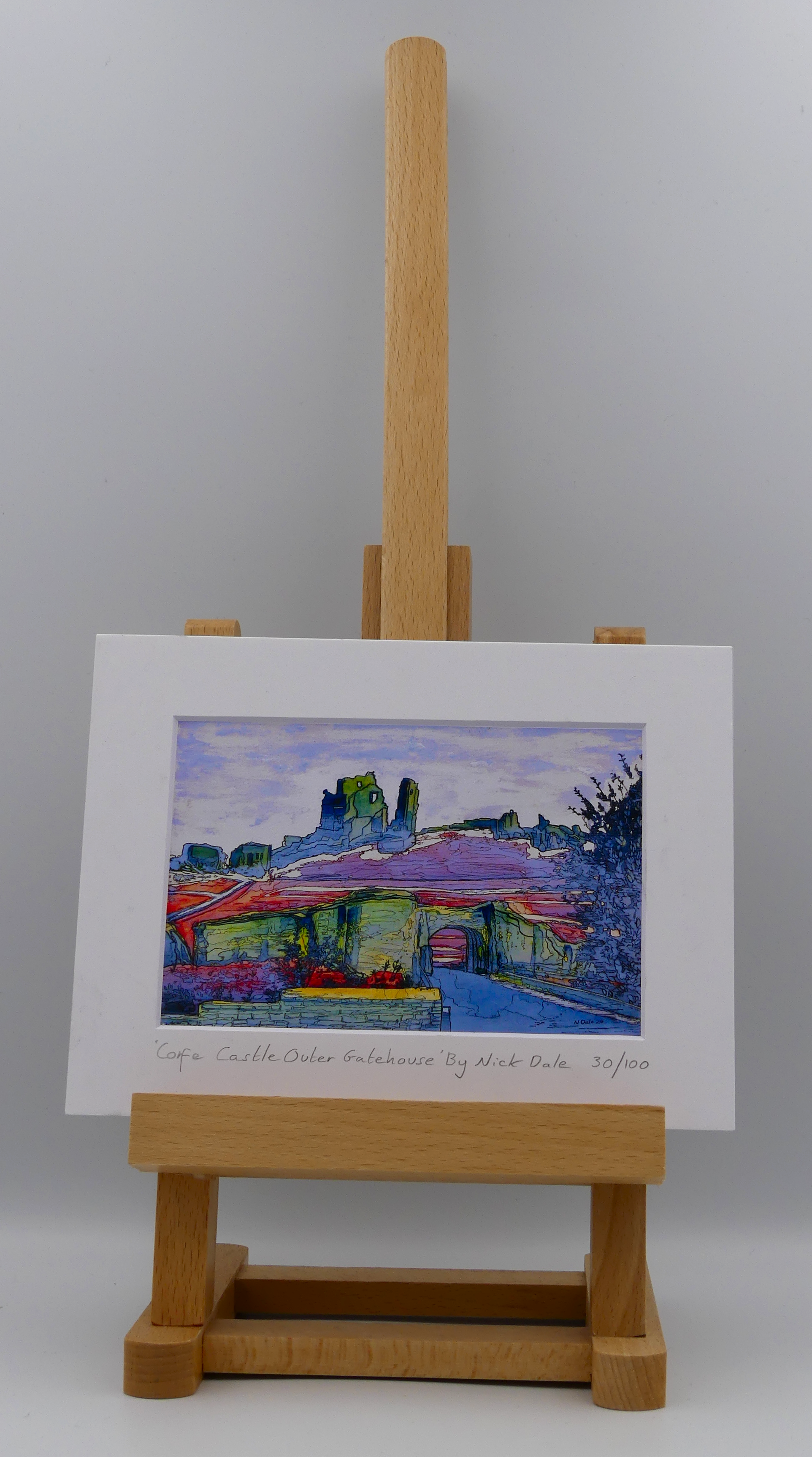 Image of a Small Art Print featuring a copy of the painting 'Corfe Castle Outer Gatehouse' by Nick Dale. The print depicts the slighted outer gatehouse of Corfe Castle in the village of Corfe Castle, Dorset. Beyond it can be seen the castle's south-west gatehouse and keep on top of the central hilltop. The print is situated on a small easel.