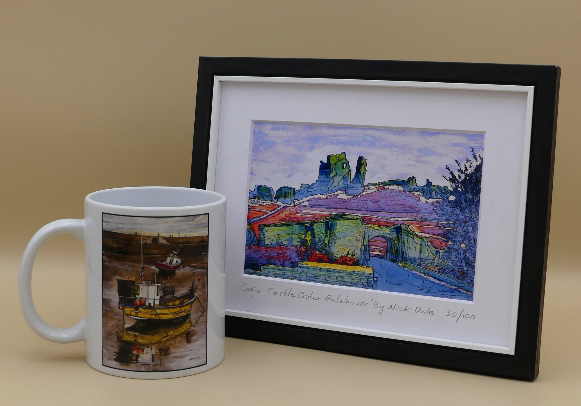 Image of a Small Art Print featuring a copy of the painting 'Corfe Castle Outer Gatehouse' by Nick Dale. The print depicts the slighted outer gatehouse of Corfe Castle in the village of Corfe Castle, Dorset. Beyond it can be seen the castle's south-west gatehouse and keep on top of the central hilltop. The print is situated in a black frame next to a mug.