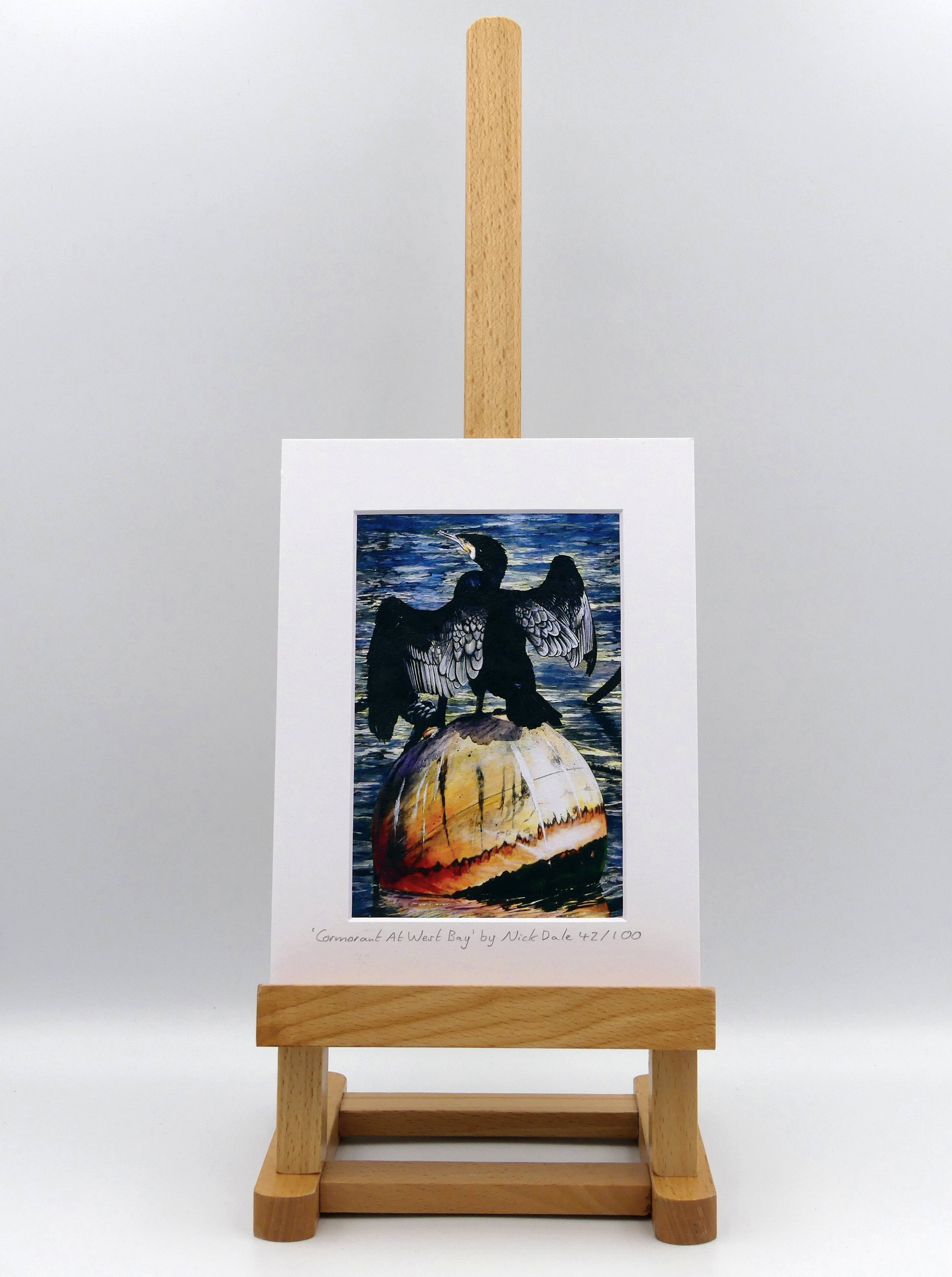 Image of Small Art Print featuring a copy of the painting 'Cormorant At West Bay' by Nick Dale. The print depicts a close-up of a cormorant sunbathing on a Mooring Buoy in West Bay's harbour in Dorset. The print is situated on a small easel.