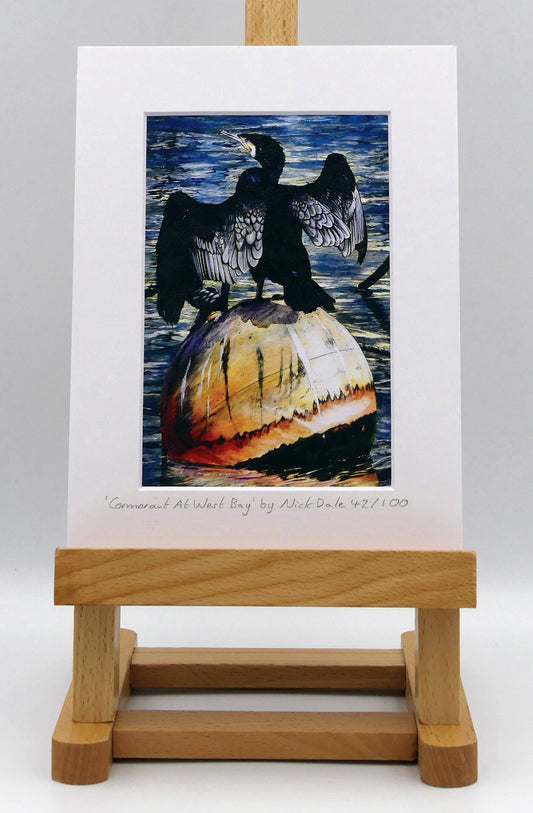 Image of Small Art Print featuring a copy of the painting 'Cormorant At West Bay' by Nick Dale. The print depicts a close-up of a cormorant sunbathing on a Mooring Buoy in West Bay's harbour in Dorset. The print is situated on a small easel.
