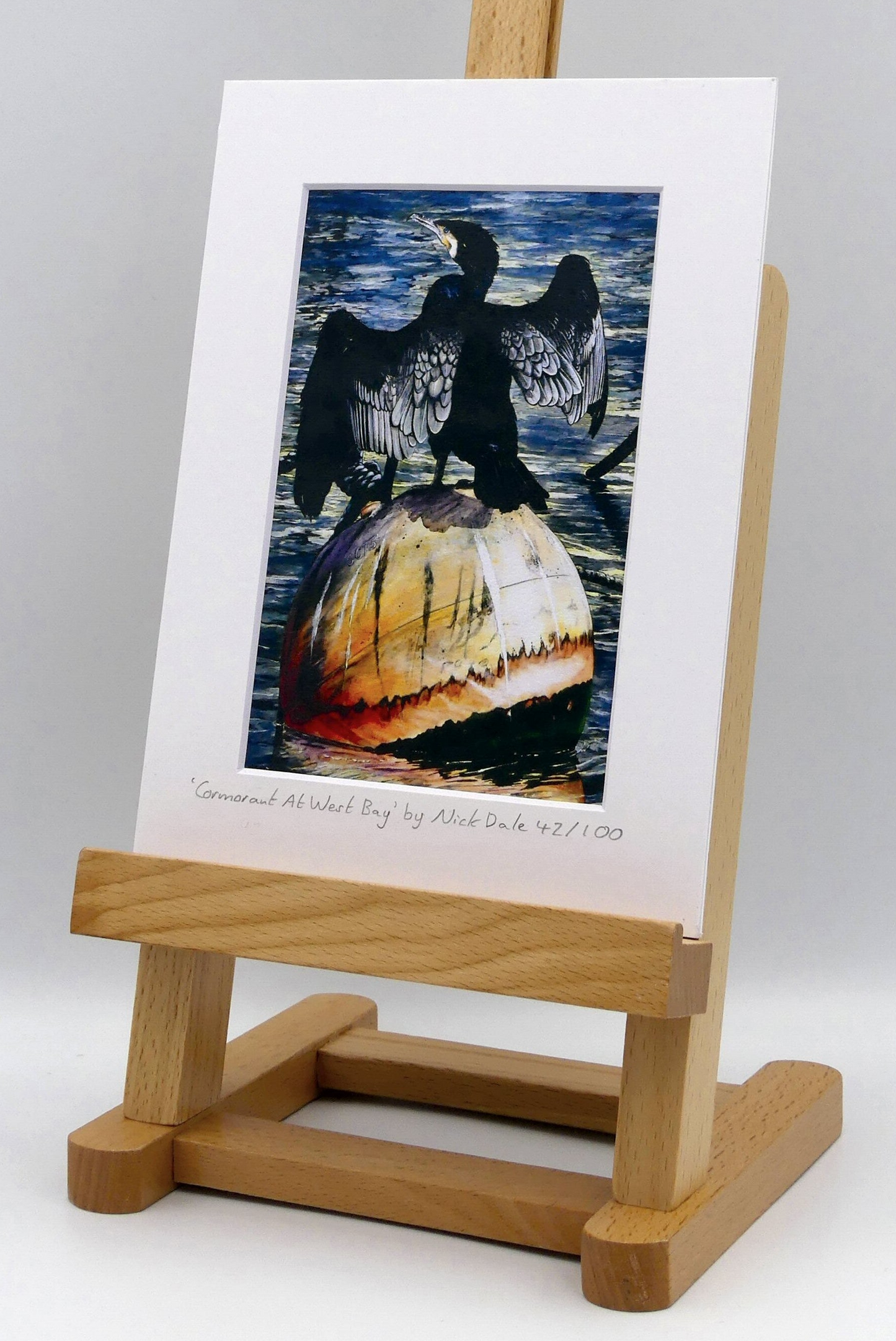 Image of Small Art Print featuring a copy of the painting 'Cormorant At West Bay' by Nick Dale. The print depicts a close-up of a cormorant sunbathing on a Mooring Buoy in West Bay's harbour in Dorset. The print is situated on a small easel.