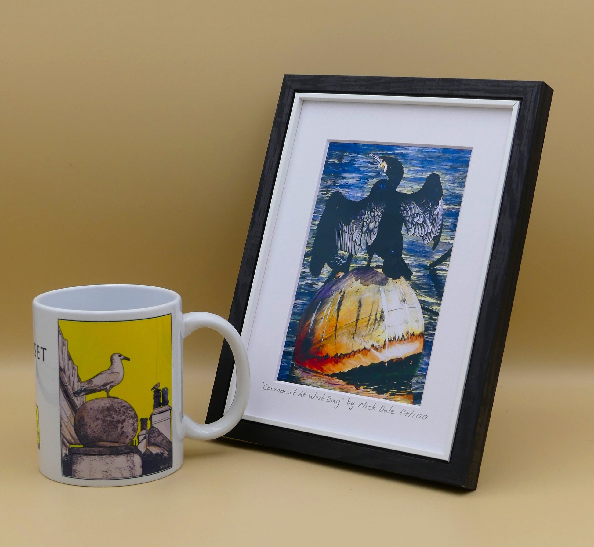 Image of Small Art Print featuring a copy of the painting 'Cormorant At West Bay' by Nick Dale. The print depicts a close-up of a cormorant sunbathing on a Mooring Buoy in West Bay's harbour in Dorset. The print is situated in a black frame next to a mug.