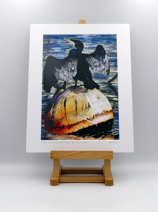 Image of Medium Art Print featuring a copy of the painting 'Cormorant At West Bay' by Nick Dale. The print depicts a close-up of a cormorant sunbathing on a Mooring Buoy in West Bay's harbour in Dorset. The print is situated on a small easel.