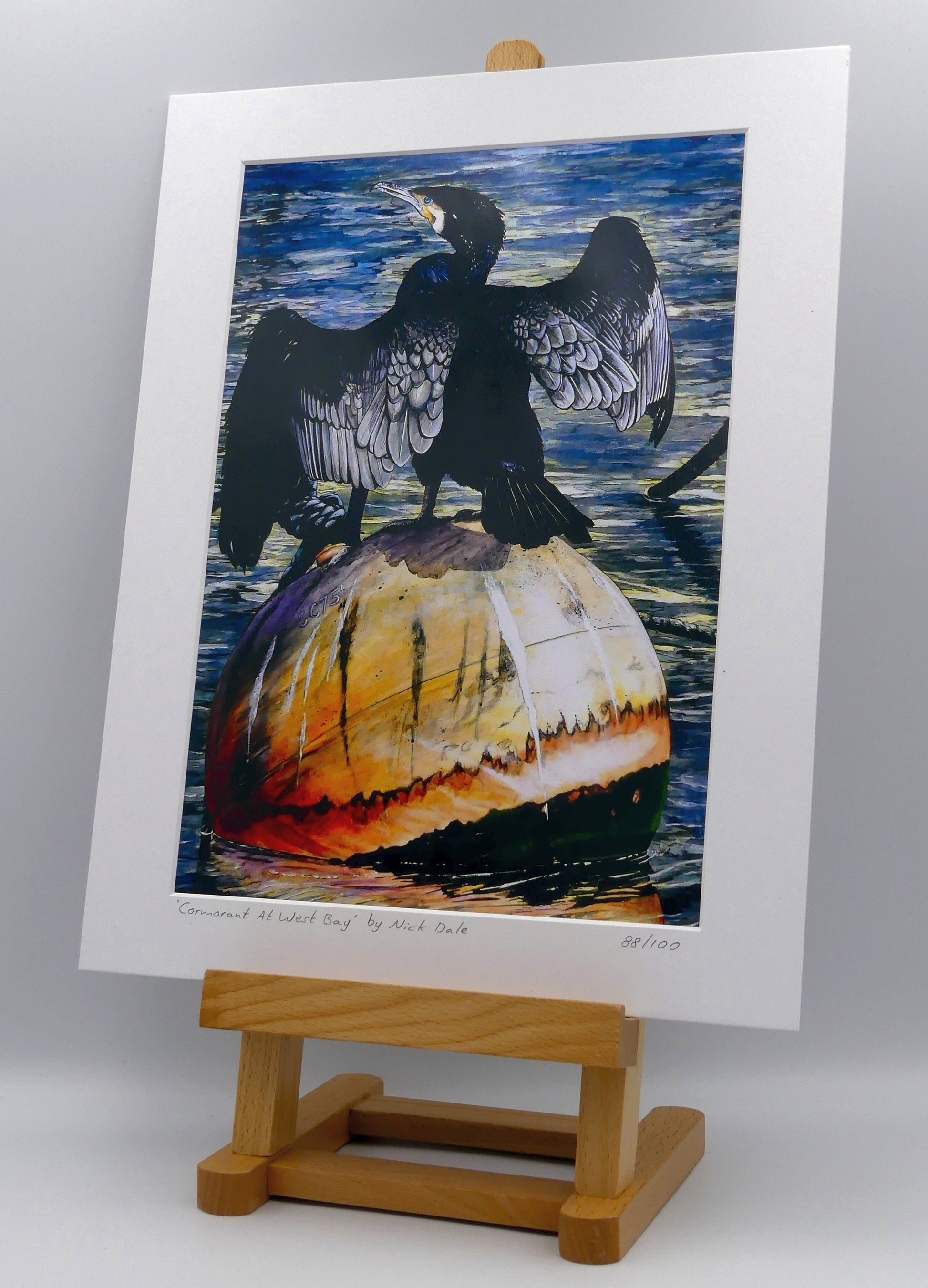 Image of Medium Art Print featuring a copy of the painting 'Cormorant At West Bay' by Nick Dale. The print depicts a close-up of a cormorant sunbathing on a Mooring Buoy in West Bay's harbour in Dorset. The print is situated on a small easel.