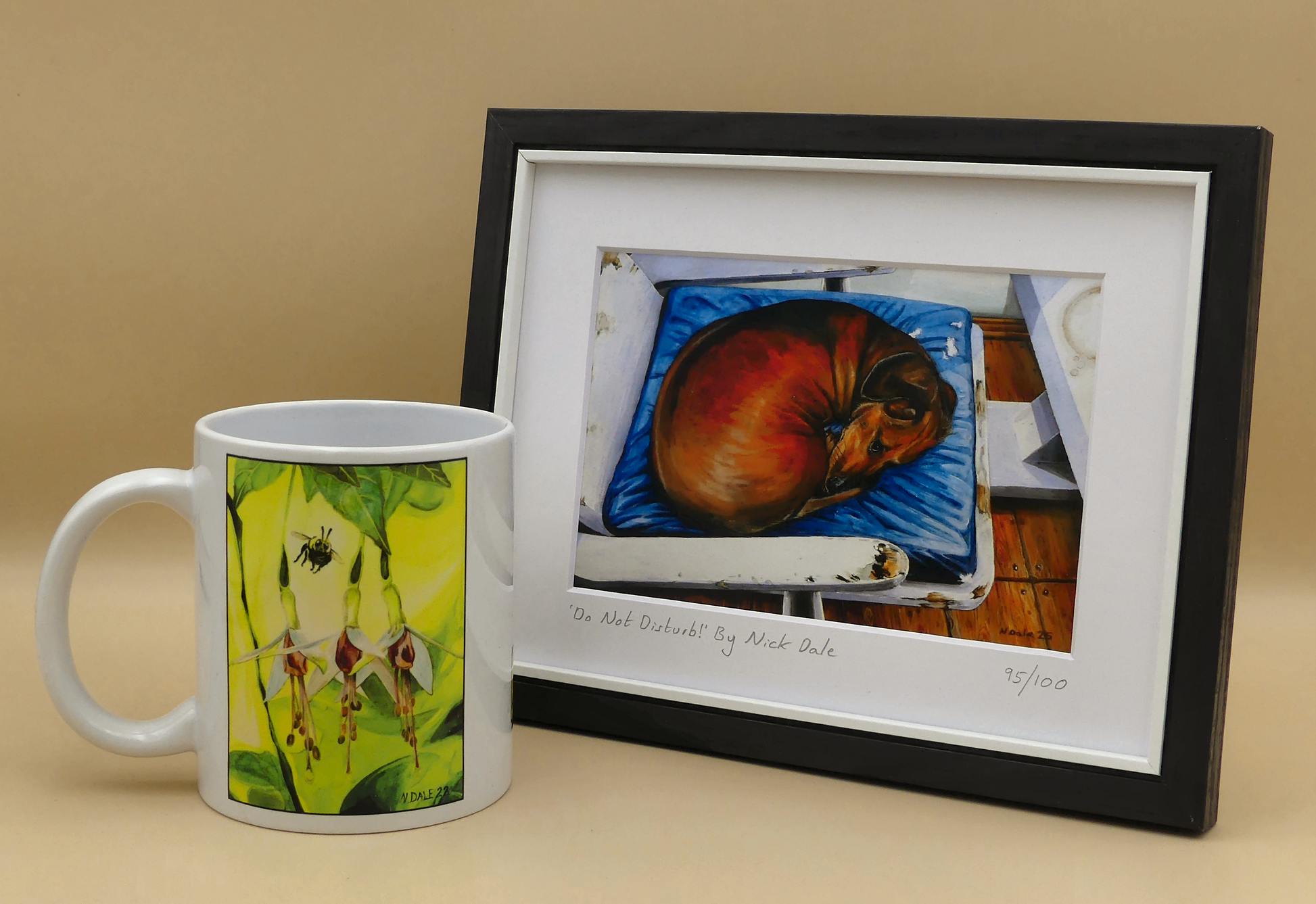 Image of a Small Art Print featuring a copy of the painting 'Do Not Disturb' by Nick Dale. The print depicts a miniature dachshund dog curled up on a favourite seat. Its gaze clearly indicates that it is not keen on being evicted from its current location and slumbering state. The print is situated in a black frame next to a mug.