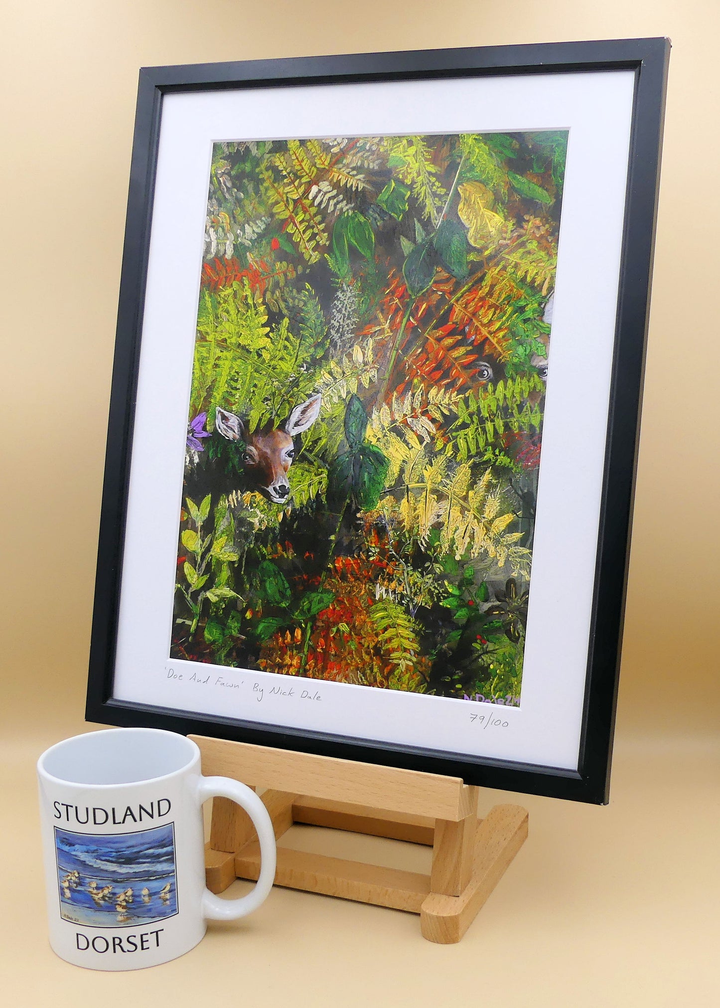 Image of a Medium Art Print featuring a copy of the painting 'Doe And Fawn' by Nick Dale. The print depicts a Row Deer doe and fawn hiding in the dense undergrowth with only their heads visible. The print is situated on a small easel in a black frame next to a mug.