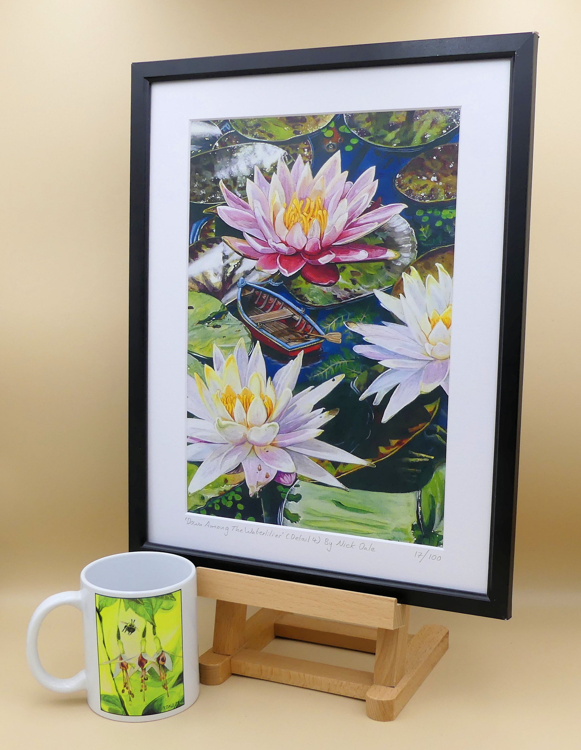 Image of a Medium Art Print featuring a copy of a detail from the painting 'Down Among The Water Lilies' by Nick Dale. The print depicts the part of the original painting showing the rowing boat and a close-up view of the water lilies at Bennetts Water Gardens in Weymouth, with the twist of the boat being shrunk down to lilliputian dimensions. The print is situated on a small easel in a black frame next to a mug.