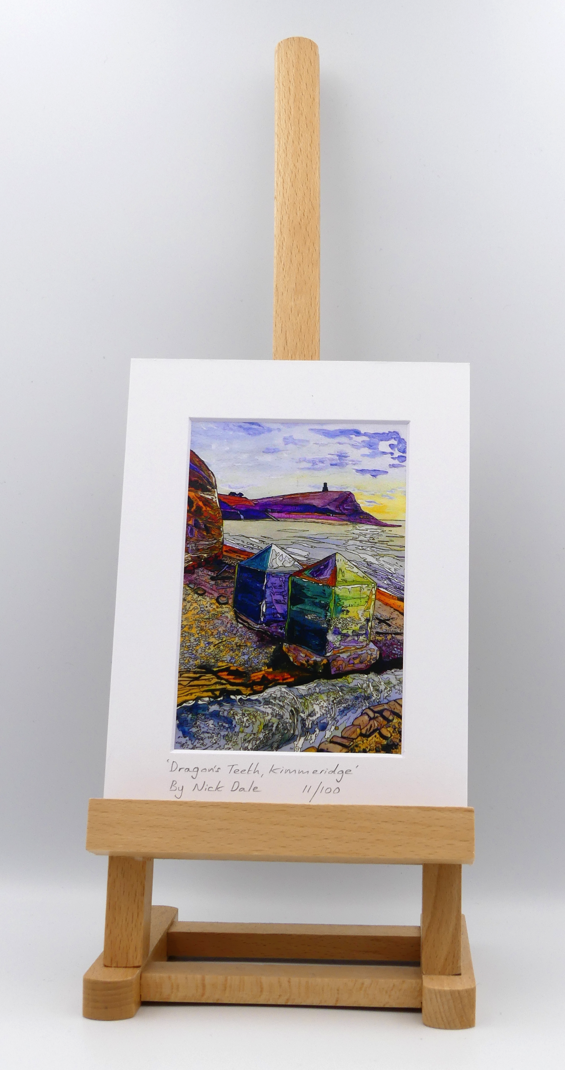 Image of a Small Art Print featuring a copy of the painting 'Dragon's Teeth, Kimmeridge' by Nick Dale. The scene depicts two surviving dragon's teeth anti-tank defences on the beach at Kimmeridge Bay, Dorset. Beyond them can be seen Clavell Tower on the cliff top. The Gaulter Gap forms a relatively easy route off the beach to the cliffs above for any invading force so it was blocked with dragon's teeth and a concrete pillbox during World War II. The print is situated on a small easel.