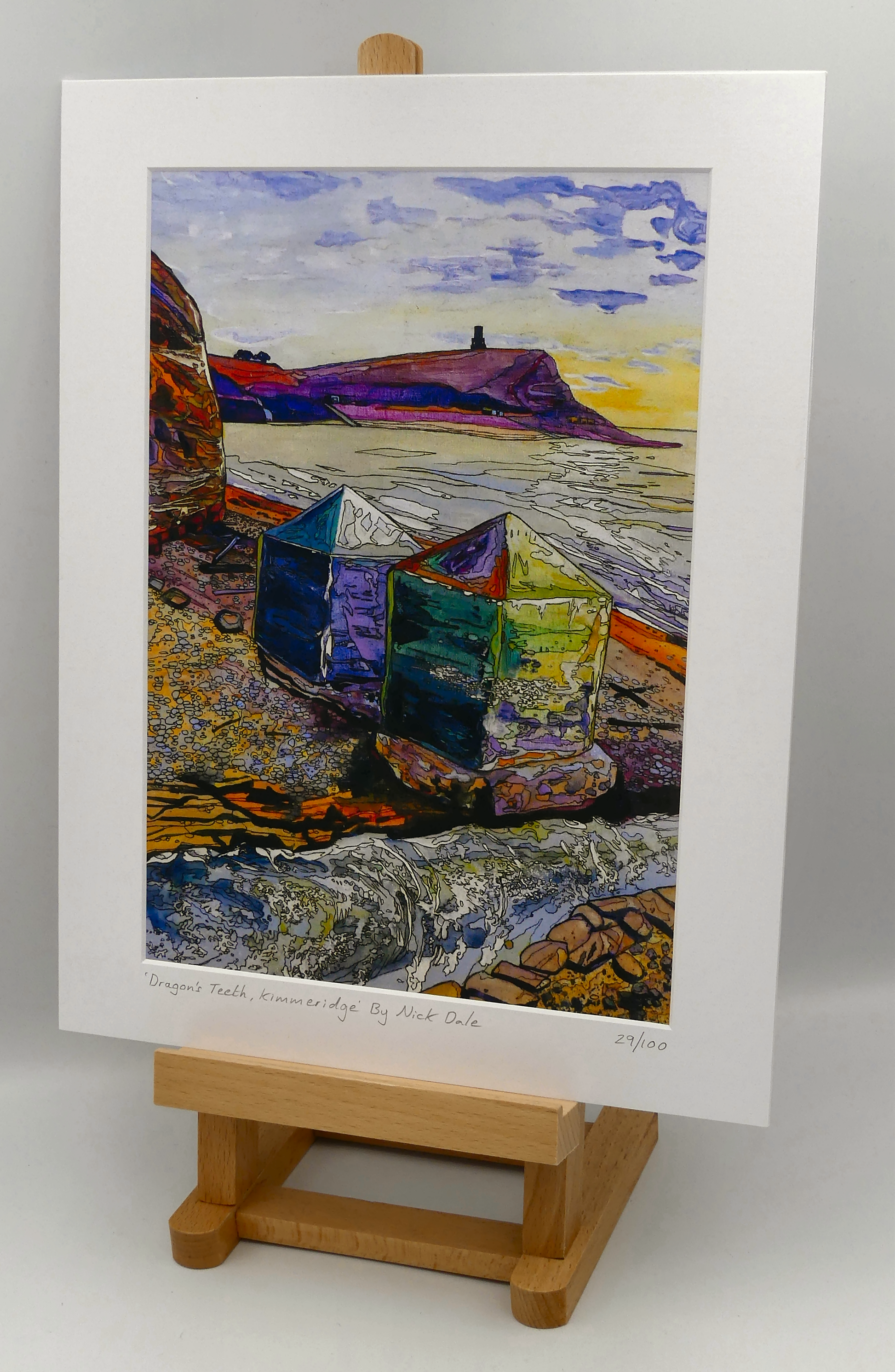 Image of a Medium Art Print featuring a copy of the painting 'Dragon's Teeth, Kimmeridge' by Nick Dale. The scene depicts two surviving dragon's teeth anti-tank defences on the beach at Kimmeridge Bay, Dorset. Beyond them can be seen Clavell Tower on the cliff top. The Gaulter Gap forms a relatively easy route off the beach to the cliffs above for any invading force so it was blocked with dragon's teeth and a concrete pillbox during World War II. The print is situated on a small easel.