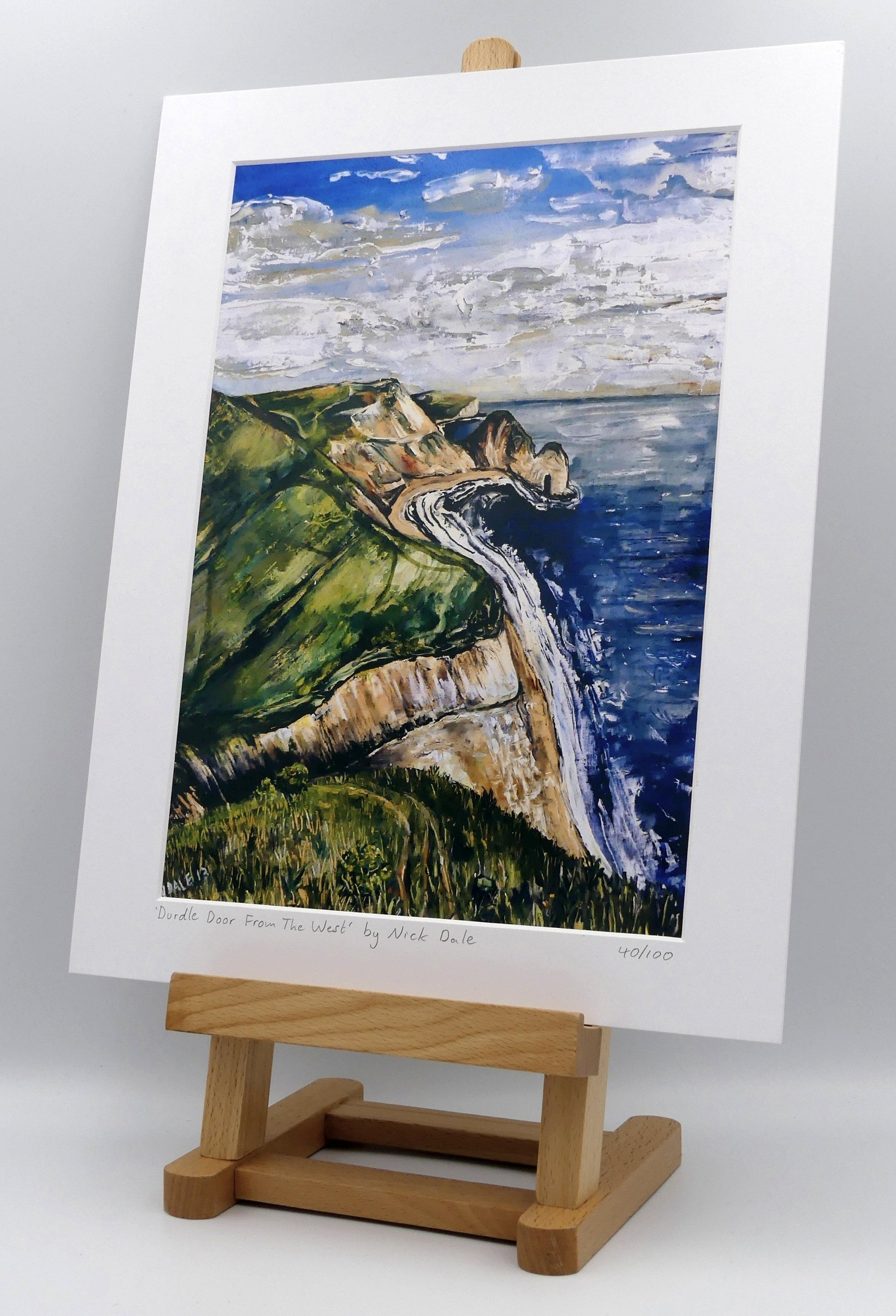 Image of a Medium Art Print featuring a copy of the painting 'Durdle Door From The West:' by Nick Dale. The print depicts a view along the Jurassic Coast to the famous Dorset limestone arch landmark, as seen from the coastal path to the west. The print is situated on a small easel.