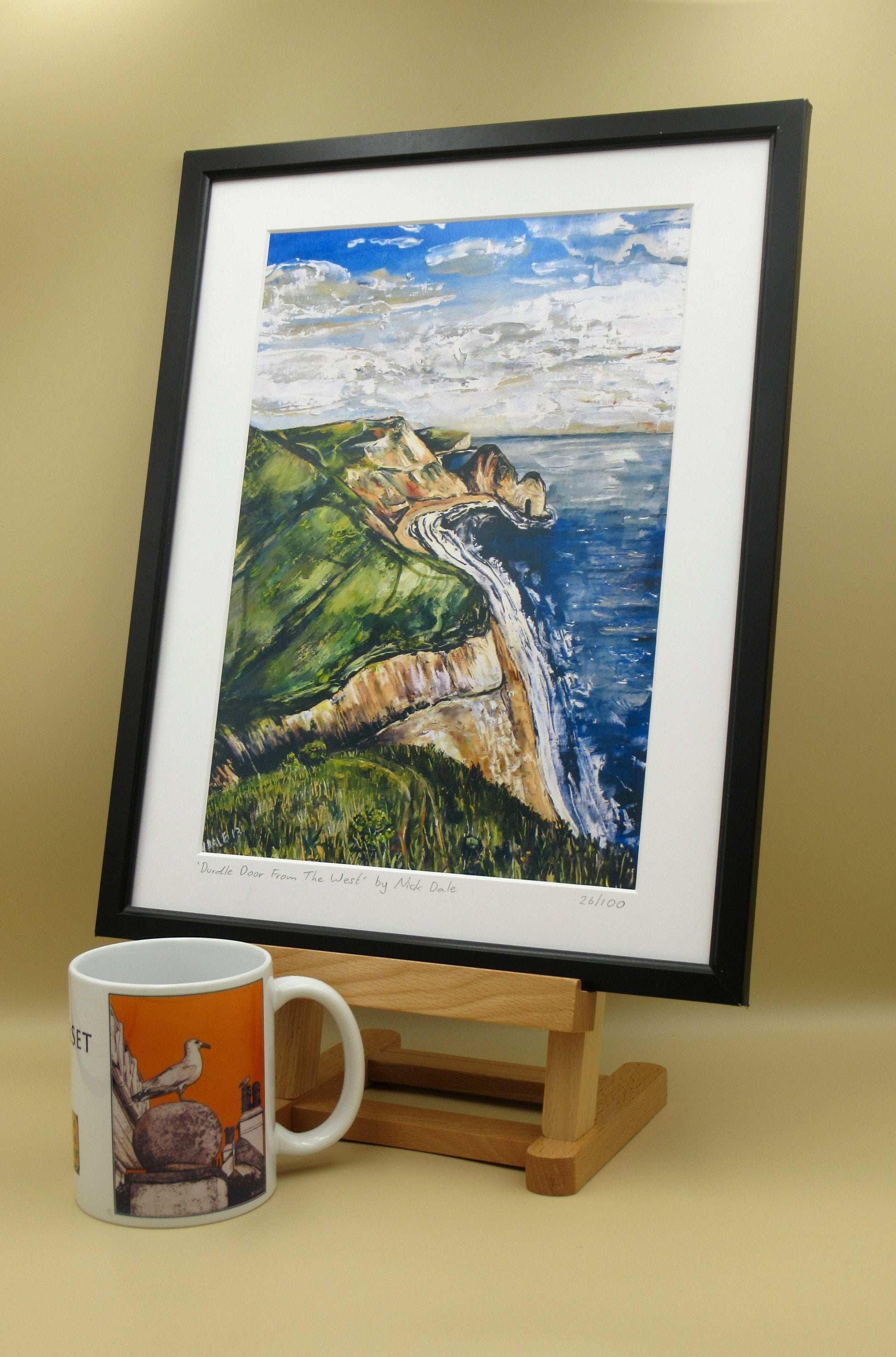 Image of a Medium Art Print featuring a copy of the painting 'Durdle Door From The West:' by Nick Dale. The print depicts a view along the Jurassic Coast to the famous Dorset limestone arch landmark, as seen from the coastal path to the west. The print is situated in a black frame next to a mug.