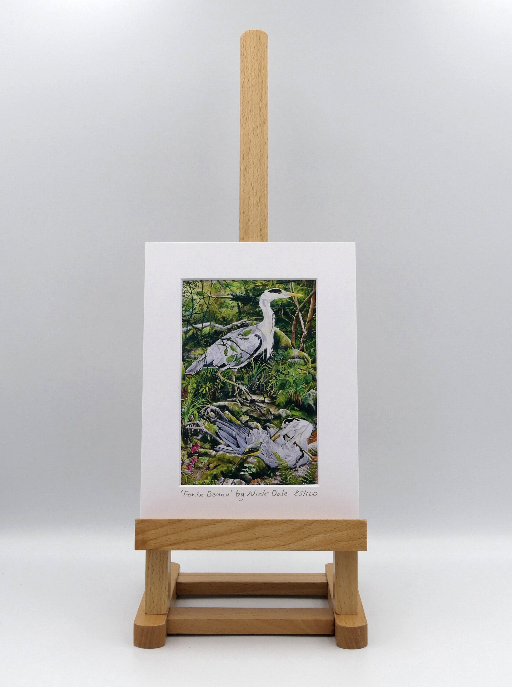 Image of a Small Art Print featuring a copy of the painting 'Fenix Bennu' by Nick Dale. The print depicts 2 grey herons (1 living & 1 dead) & is named after the Egyptian god Bennu; which may be the origins of the phoenix legend. 'Fenix' is Anglo-Saxon for phoenix & the scene has been transposed to a river in Devon. The blood of the dead heron is smoking where it drips on the ground; suggesting that this is indeed a phoenix. The print is situated on a small easel.