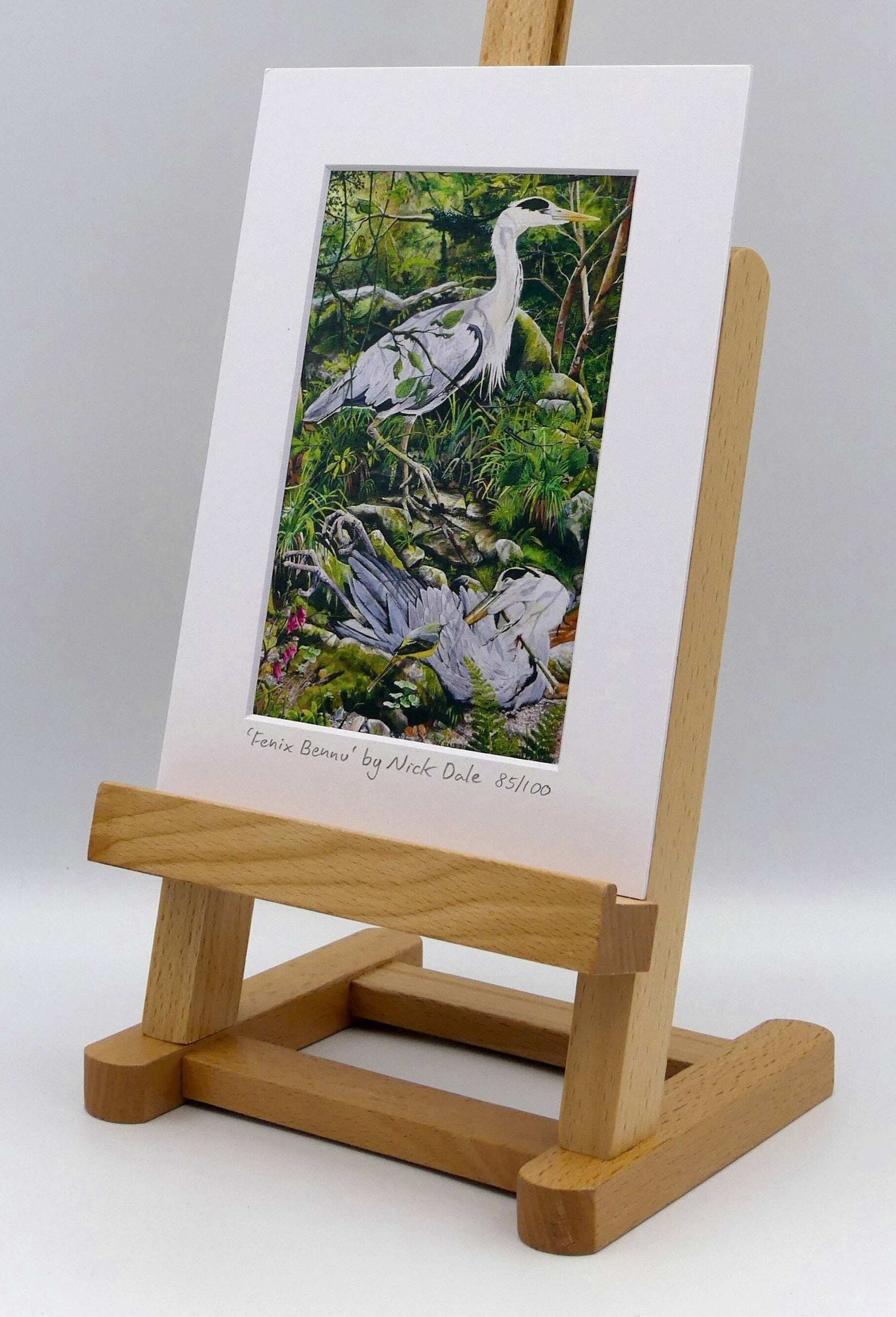 Image of a Small Art Print featuring a copy of the painting 'Fenix Bennu' by Nick Dale. The print depicts 2 grey herons (1 living & 1 dead) & is named after the Egyptian god Bennu; which may be the origins of the phoenix legend. 'Fenix' is Anglo-Saxon for phoenix & the scene has been transposed to a river in Devon. The blood of the dead heron is smoking where it drips on the ground; suggesting that this is indeed a phoenix. The print is situated on a small easel.