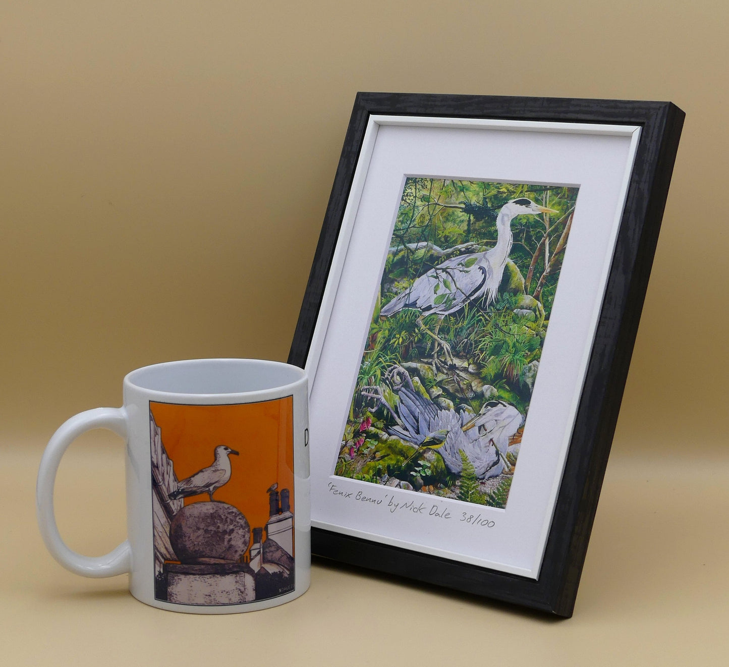 Image of a Small Art Print featuring a copy of the painting 'Fenix Bennu' by Nick Dale. The print depicts 2 grey herons (1 living & 1 dead) & is named after the Egyptian god Bennu; which may be the origins of the phoenix legend. 'Fenix' is Anglo-Saxon for phoenix & the scene has been transposed to a river in Devon. The blood of the dead heron is smoking where it drips on the ground; suggesting that this is indeed a phoenix. The print is situated in a black frame next to a mug.