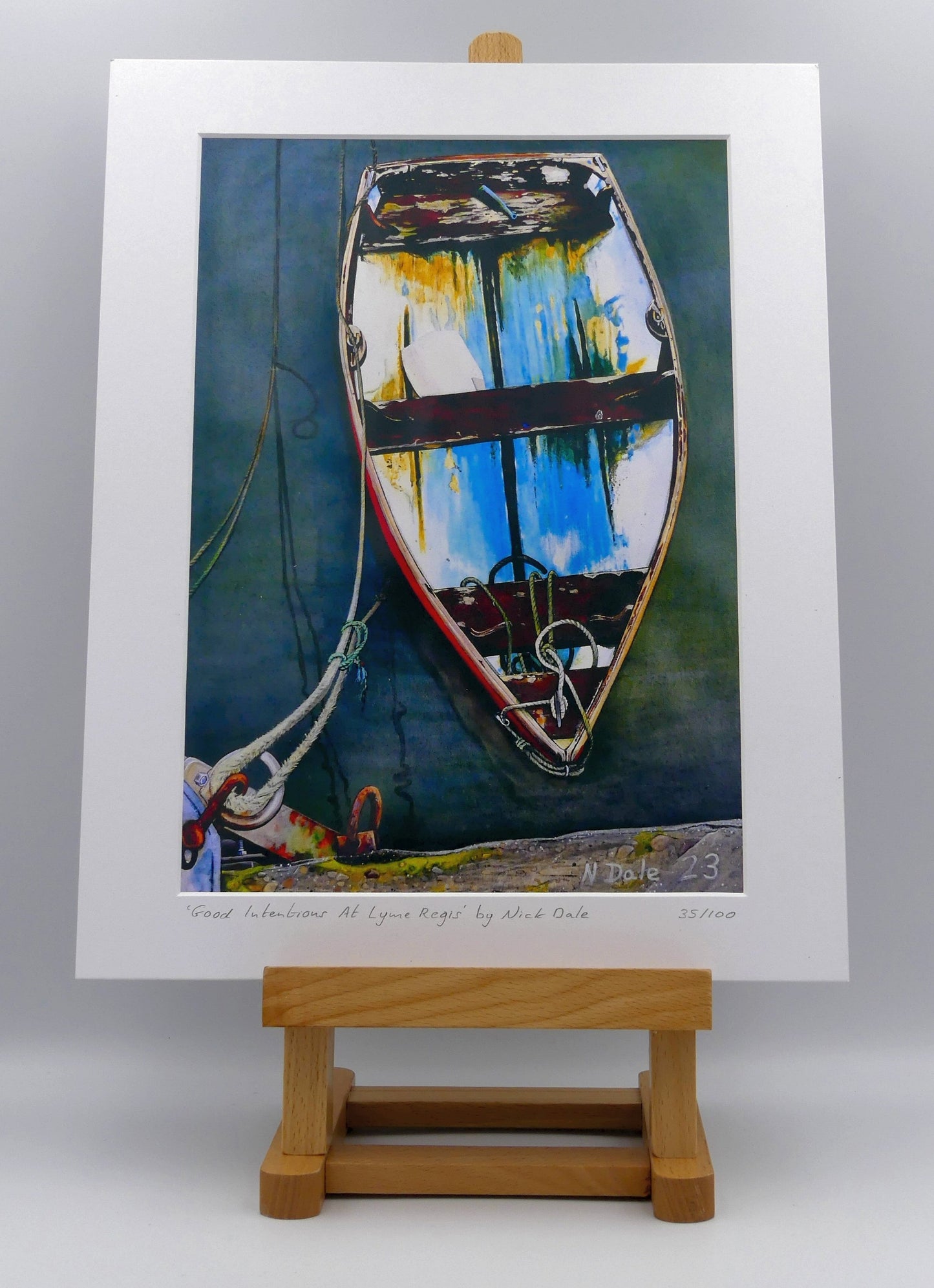 Image of a Medium Art Print featuring a copy of the painting 'Good Intentions at Lyme Regis' by Nick Dale. The print depicts a small wooden rowing boat tied up in Lyme Regis harbour, Dorset. The print is situated on a small easel.