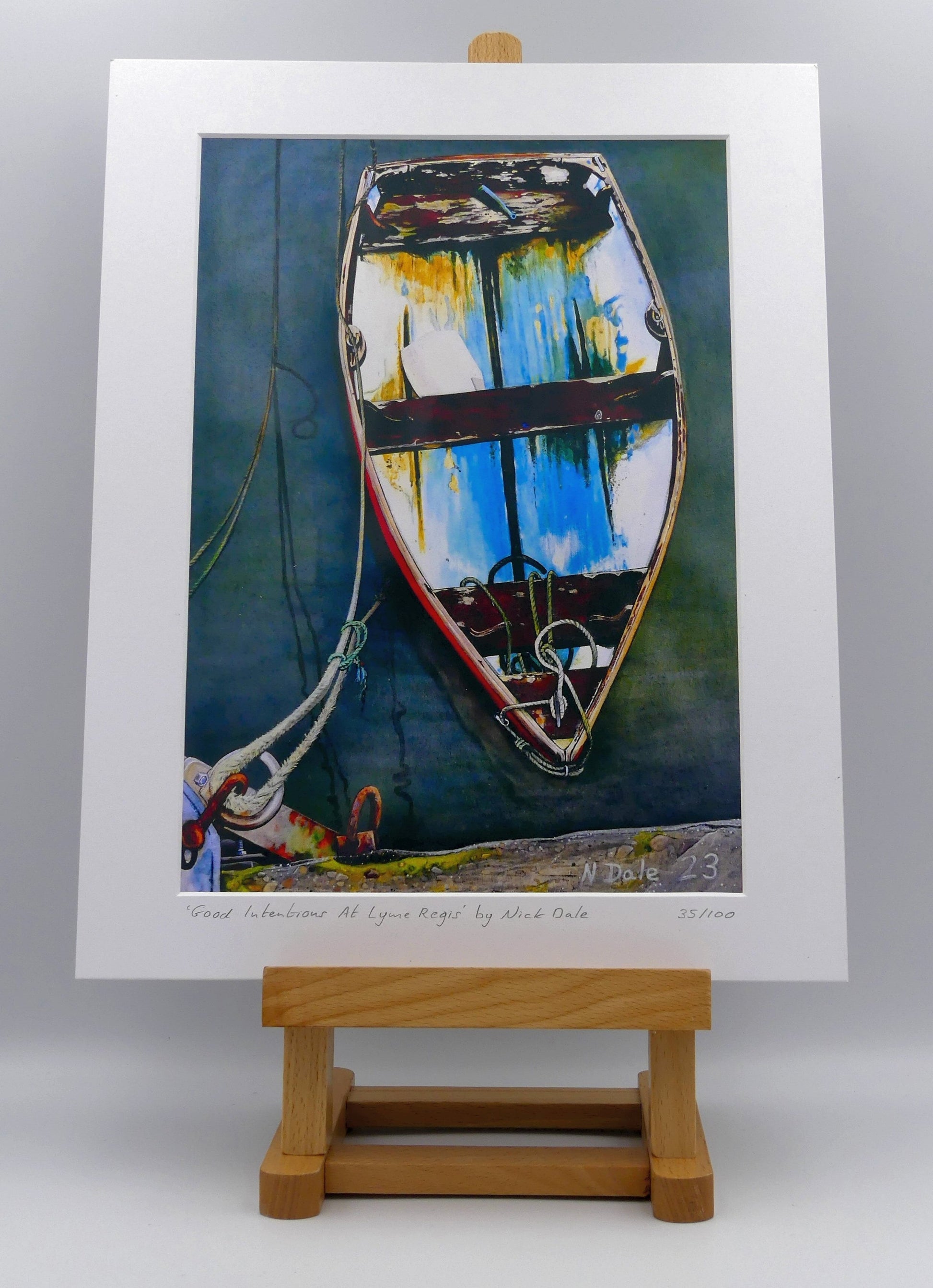 Image of a Medium Art Print featuring a copy of the painting 'Good Intentions at Lyme Regis' by Nick Dale. The print depicts a small wooden rowing boat tied up in Lyme Regis harbour, Dorset. The print is situated on a small easel.