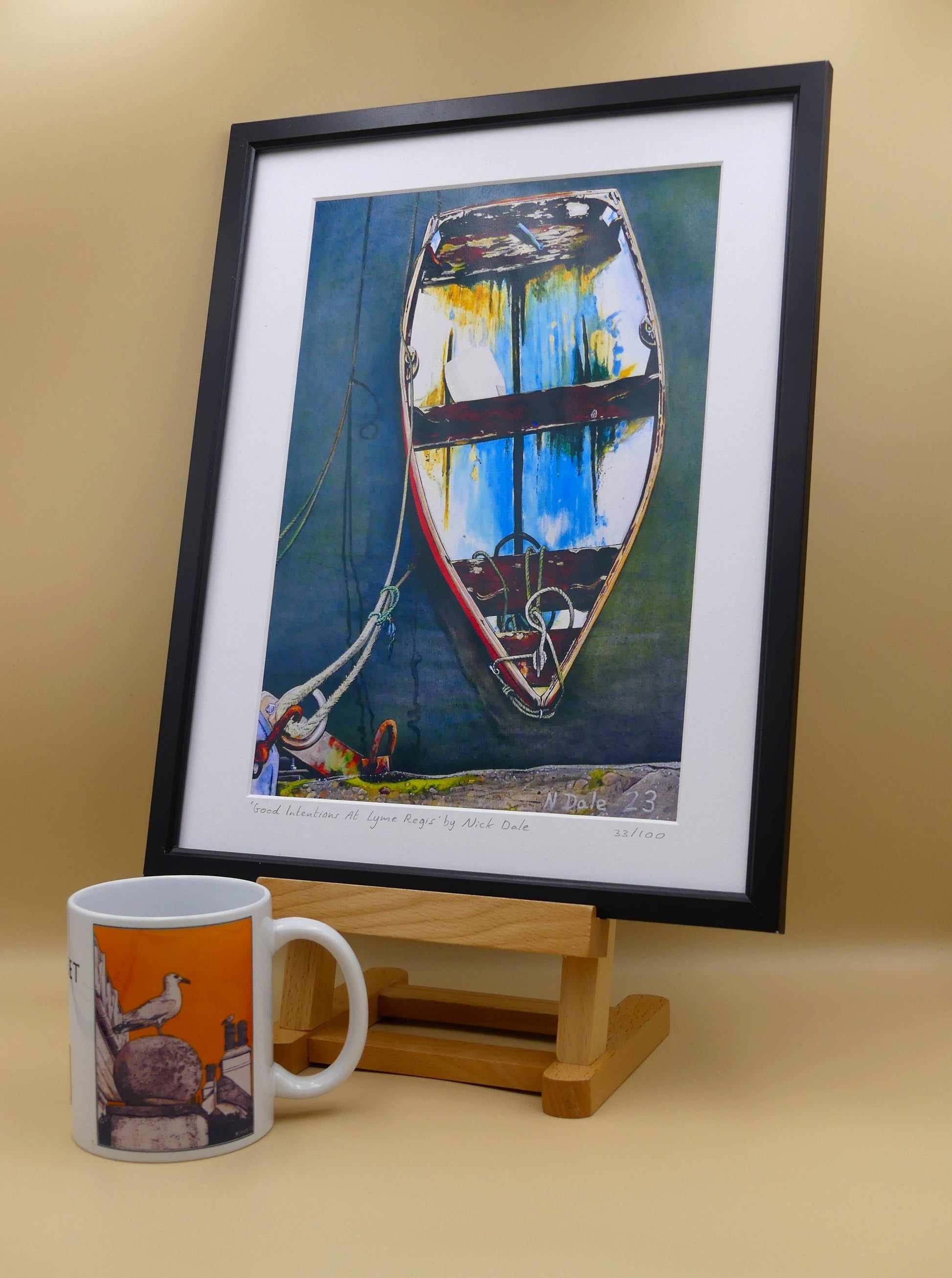 Image of a Medium Art Print featuring a copy of the painting 'Good Intentions at Lyme Regis' by Nick Dale. The print depicts a small wooden rowing boat tied up in Lyme Regis harbour, Dorset. The print is situated in a black frame next to a mug.