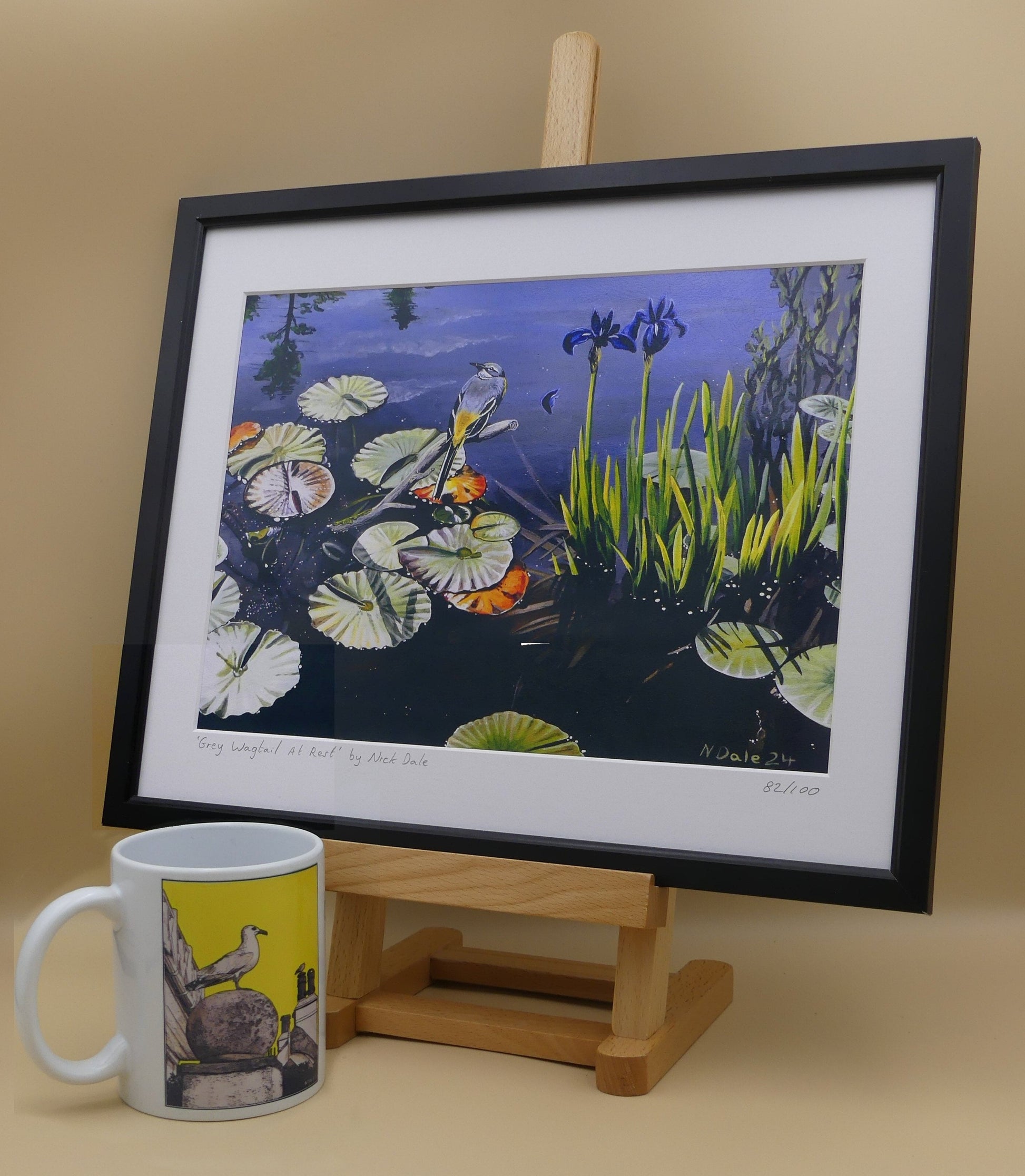 Image of Medium Art Print featuring a copy of the painting 'Grey Wagtail At Rest' by Nick Dale. The print depicts a grey wagtail bird at rest, perched on top of a submerged stick above a pond. Whilst the grey wagtail was observed at Athelhampton House in Dorset the pond's vegetation is more closely based on a pond at Knightshayes Court in Devon. The print is situated in a black frame next to a mug.