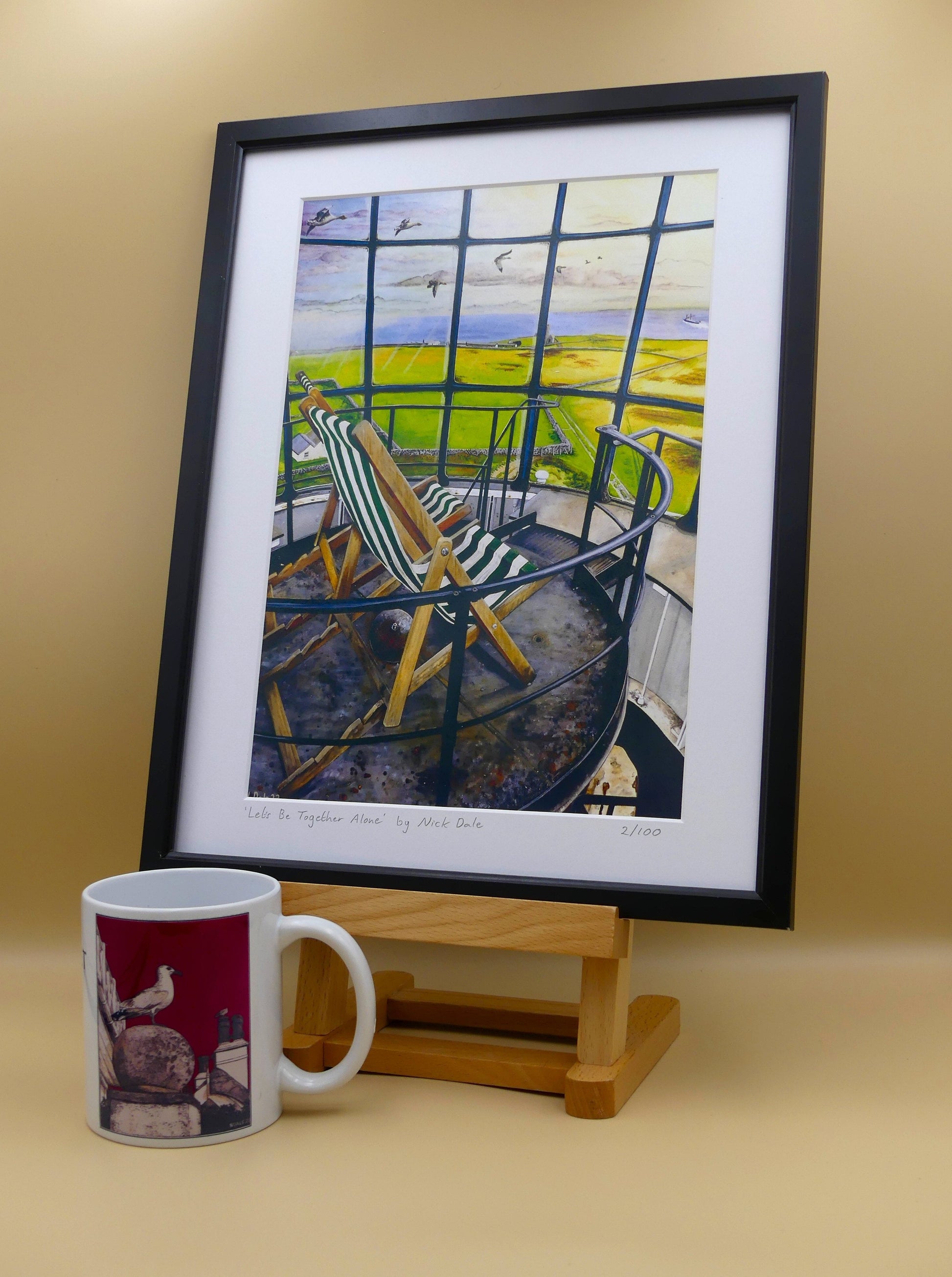 Image of a Medium Art Print featuring a copy of the painting 'Let's Be Together Alone' by Nick Dale. The print depicts the view from the lantern room of the Old Light lighthouse on Beacon Hill, on the island of Lundy, Devon. The print is situated in a black frame next to a mug.