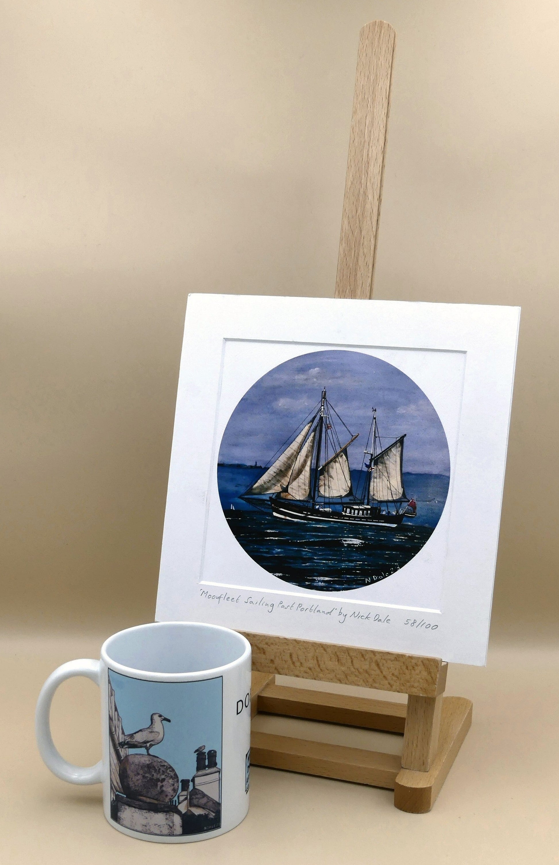 Image of Square Medium Art Print featuring a copy of the painting 'Moonfleet Sailing Past Portland' by Nick Dale. The print depicts the tall ship Moonfleet sailing past the western coast of Portland, Dorset. As observed from Ringstead Beach. The print is situated on a small easel next to a mug.