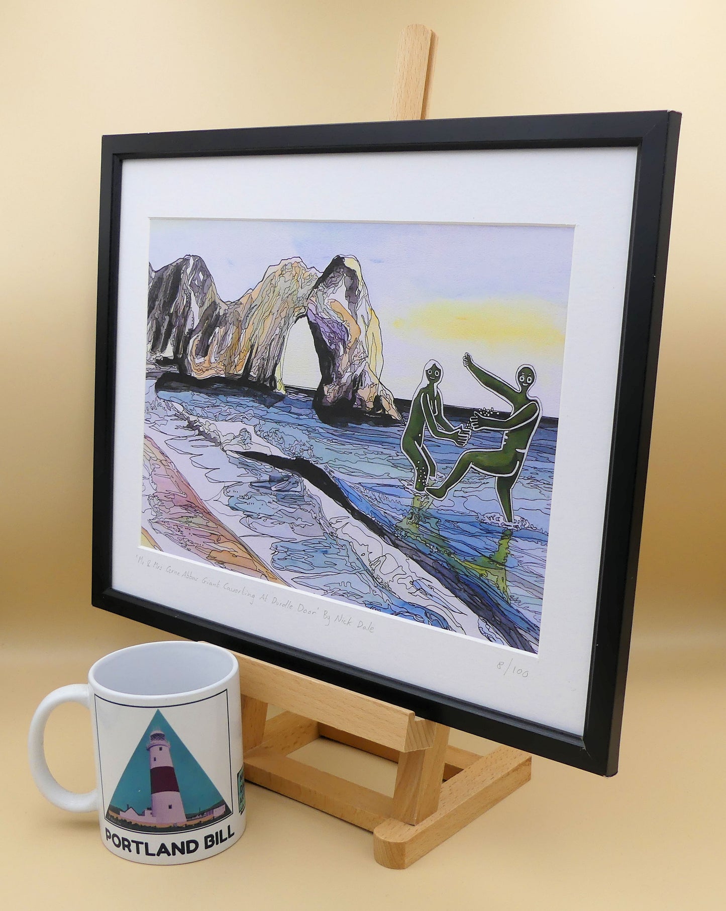 Image of a Medium Art Print featuring a copy of the painting 'Mr & Mrs Cerne Abbas Giant Cavorting At Durdle Door' by Nick Dale. The scene imagines the future Mr & Mrs Cerne Abbas Giant's first date at Durdle Door; where they cavort in the water in front of the famous Dorset landmark. The print is situated on a small easel in a black frame next to a mug.
