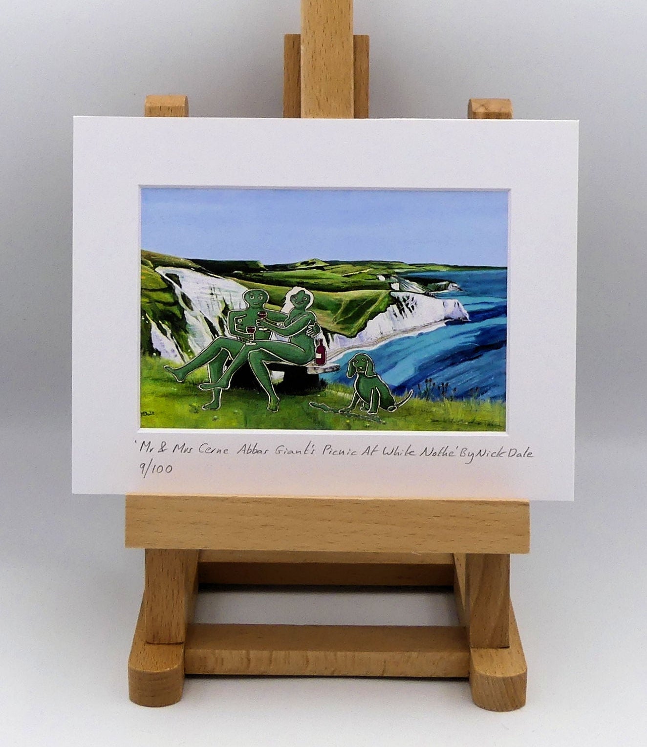 Image of a Small Art Print featuring a copy of the composite painting 'Mr & Mrs Cerne Abbas Giant's Picnic At White Nothe' by Nick Dale. The print depicts Mr & Mrs Cerne Abbas Giant having a prosecco picnic whilst on a date along the Dorset coast at the White Nothe, east of Weymouth. They are joined on the date by their cocker spaniel dog.  The print is situated on a small easel.