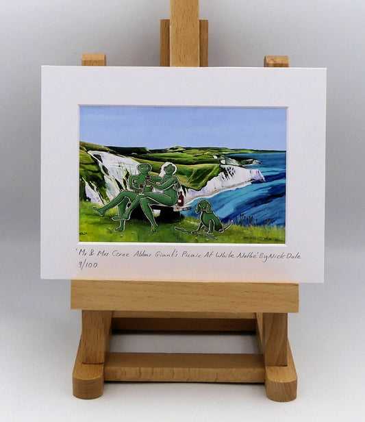 Image of a Small Art Print featuring a copy of the composite painting 'Mr & Mrs Cerne Abbas Giant's Picnic At White Nothe' by Nick Dale. The print depicts Mr & Mrs Cerne Abbas Giant having a prosecco picnic whilst on a date along the Dorset coast at the White Nothe, east of Weymouth. They are joined on the date by their cocker spaniel dog.  The print is situated on a small easel.