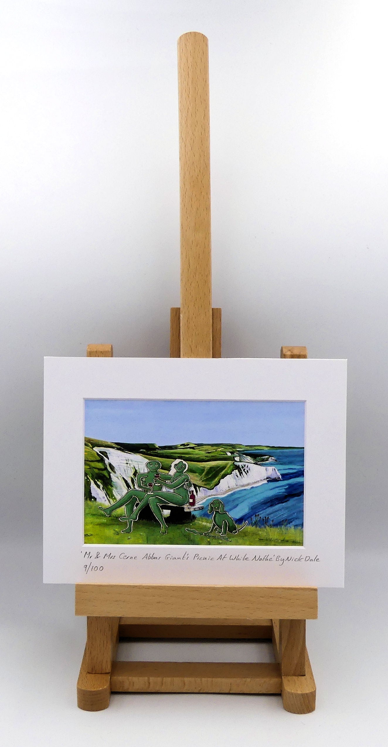 Image of a Small Art Print featuring a copy of the composite painting 'Mr & Mrs Cerne Abbas Giant's Picnic At White Nothe' by Nick Dale. The print depicts Mr & Mrs Cerne Abbas Giant having a prosecco picnic whilst on a date along the Dorset coast at the White Nothe, east of Weymouth. They are joined on the date by their cocker spaniel dog. The print is situated on a small easel.