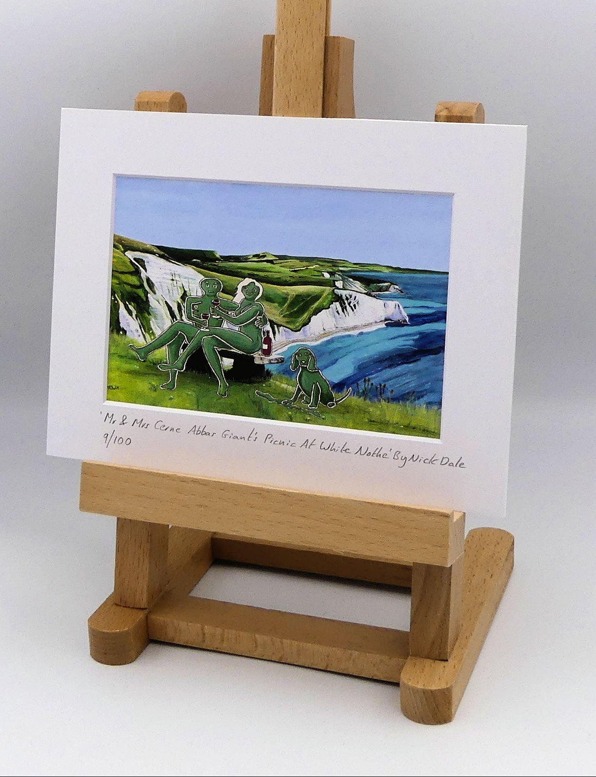 Image of a Small Art Print featuring a copy of the composite painting 'Mr & Mrs Cerne Abbas Giant's Picnic At White Nothe' by Nick Dale. The print depicts Mr & Mrs Cerne Abbas Giant having a prosecco picnic whilst on a date along the Dorset coast at the White Nothe, east of Weymouth. They are joined on the date by their cocker spaniel dog. The print is situated on a small easel.