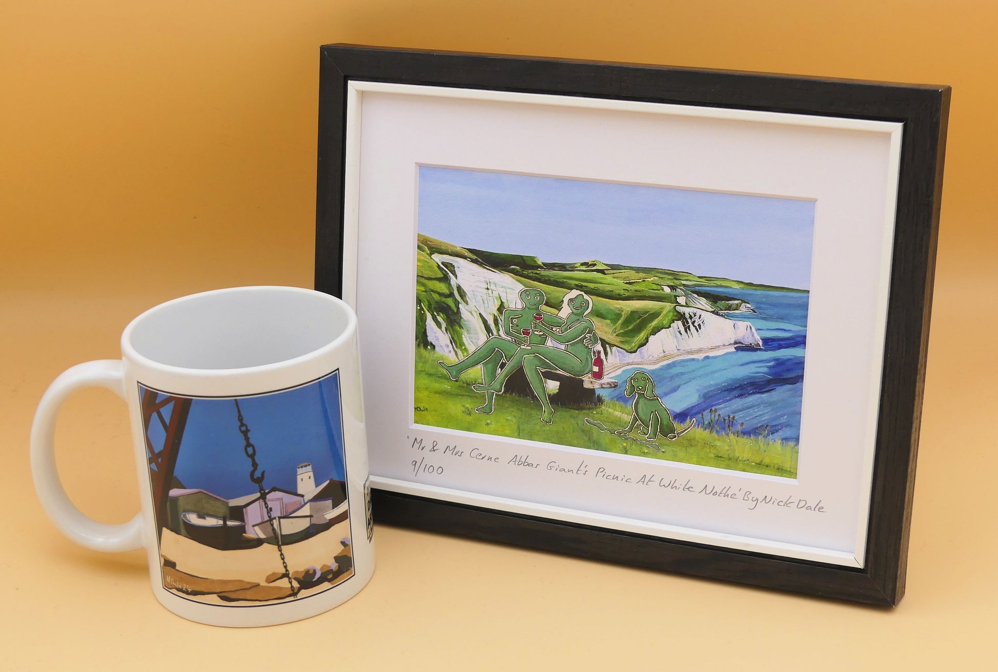 Image of a Small Art Print featuring a copy of the composite painting 'Mr & Mrs Cerne Abbas Giant's Picnic At White Nothe' by Nick Dale. The print depicts Mr & Mrs Cerne Abbas Giant having a prosecco picnic whilst on a date along the Dorset coast at the White Nothe, east of Weymouth. They are joined on the date by their cocker spaniel dog. The print is situated in a black frame next to a mug.