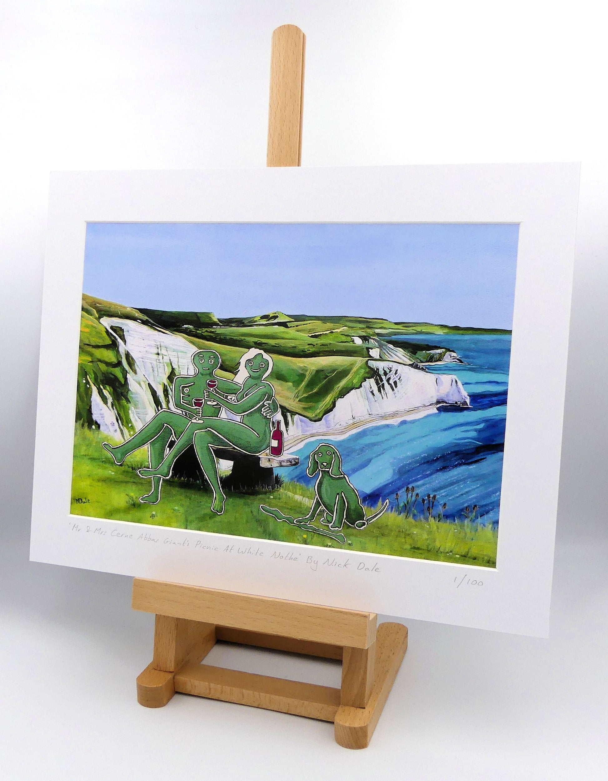 Image of a Medium Art Print featuring a copy of the composite painting 'Mr & Mrs Cerne Abbas Giant's Picnic At White Nothe' by Nick Dale. The print depicts Mr & Mrs Cerne Abbas Giant having a merlot picnic whilst on a date along the Dorset coast at the White Nothe, east of Weymouth. They are joined on the date by their cocker spaniel dog. The print is situated on a small easel.