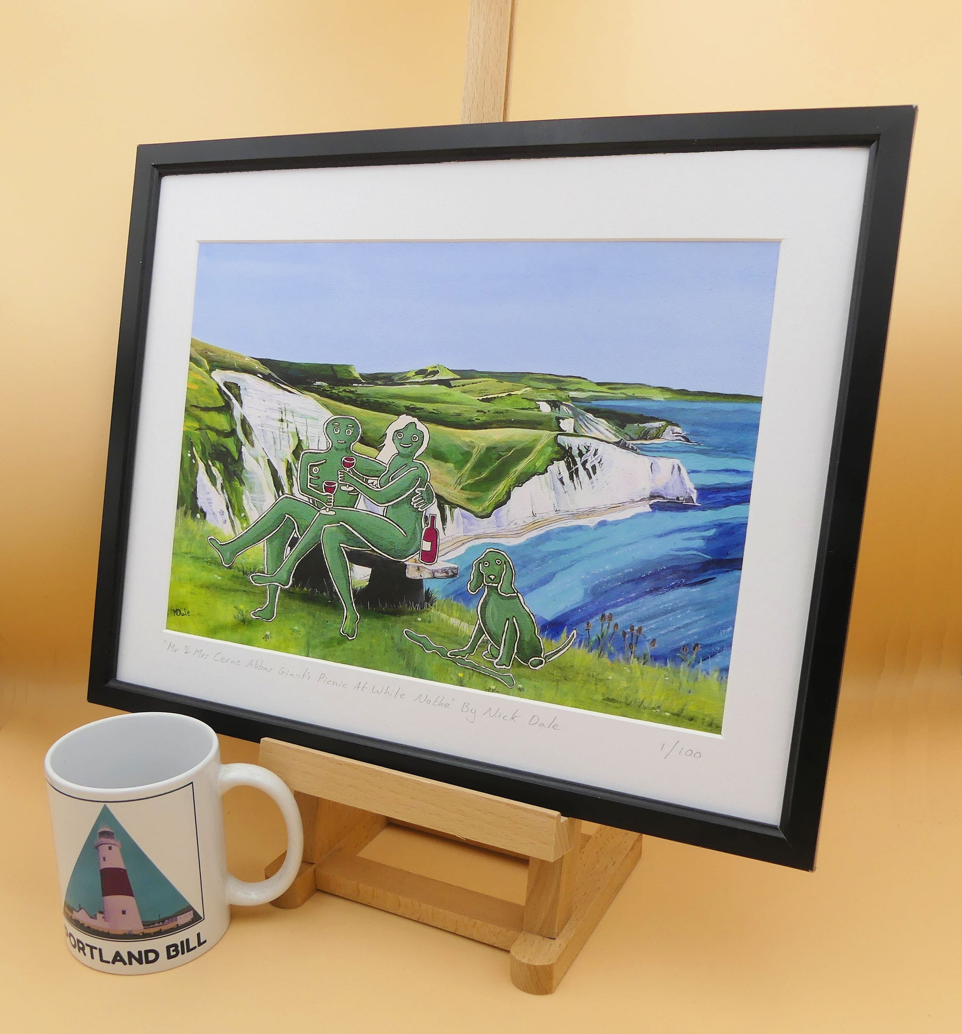 Image of a Medium Art Print featuring a copy of the composite painting 'Mr & Mrs Cerne Abbas Giant's Picnic At White Nothe' by Nick Dale. The print depicts Mr & Mrs Cerne Abbas Giant having a merlot picnic whilst on a date along the Dorset coast at the White Nothe, east of Weymouth. They are joined on the date by their cocker spaniel dog. The print is situated on a small easel in a black frame next to a mug.