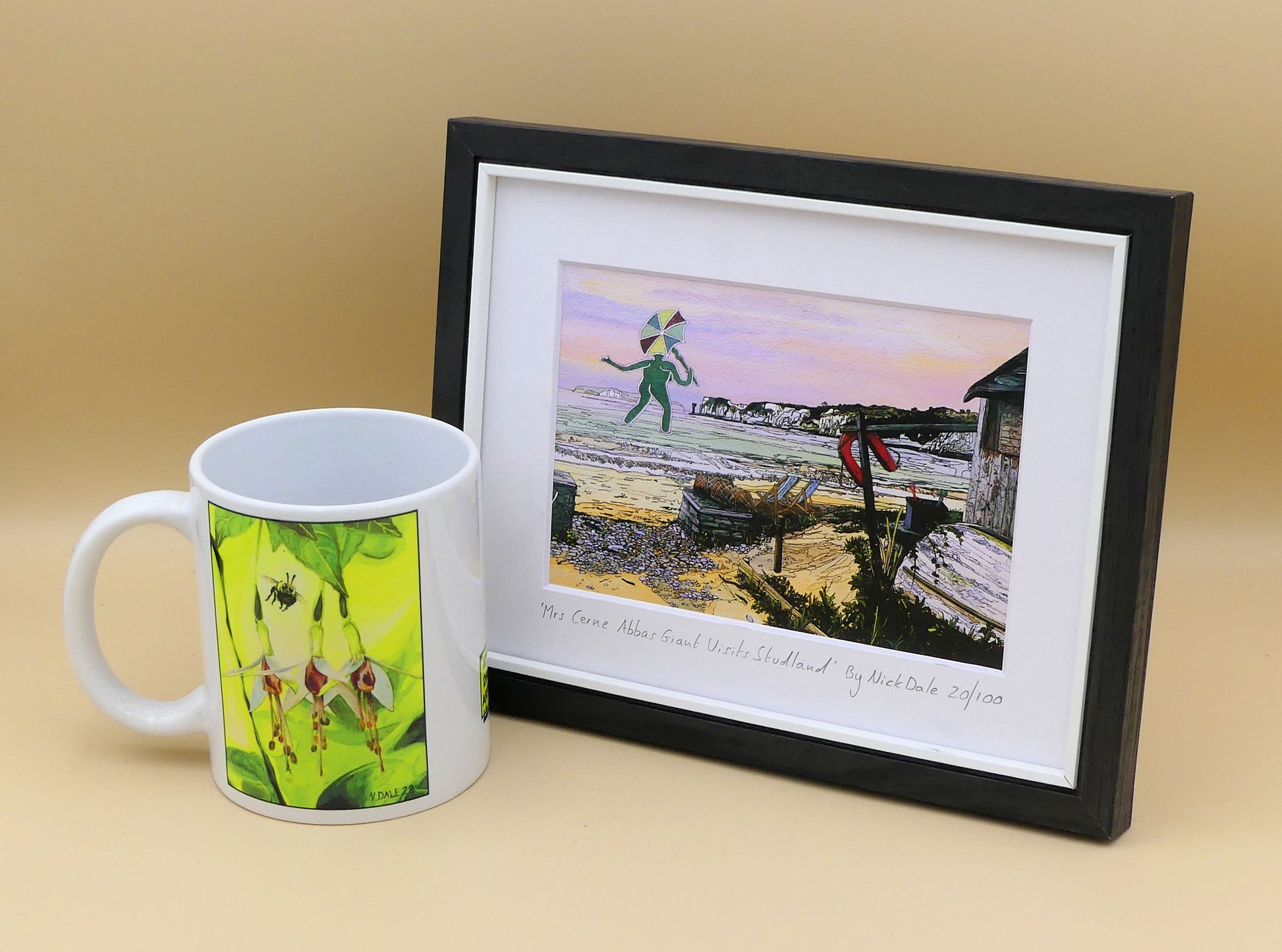 Image of a Small Art Print featuring a copy of the painting 'Mrs Cerne Abbas Giant Visits Studland' by Nick Dale. The print imagines the Cerne Abbas Giant's wife visiting the seaside at Studland Beach; with Old Harry Rocks and the Isle of Wight in the distance. The print is situated in a black frame next to a mug.